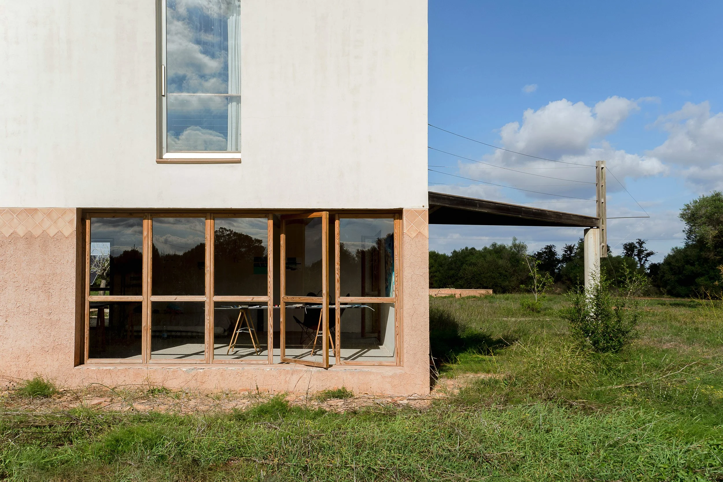 A partially constructed or renovated building with large glass windows and a small window above. The building is situated in a grassy, rural area with trees and a clear blue sky overhead.