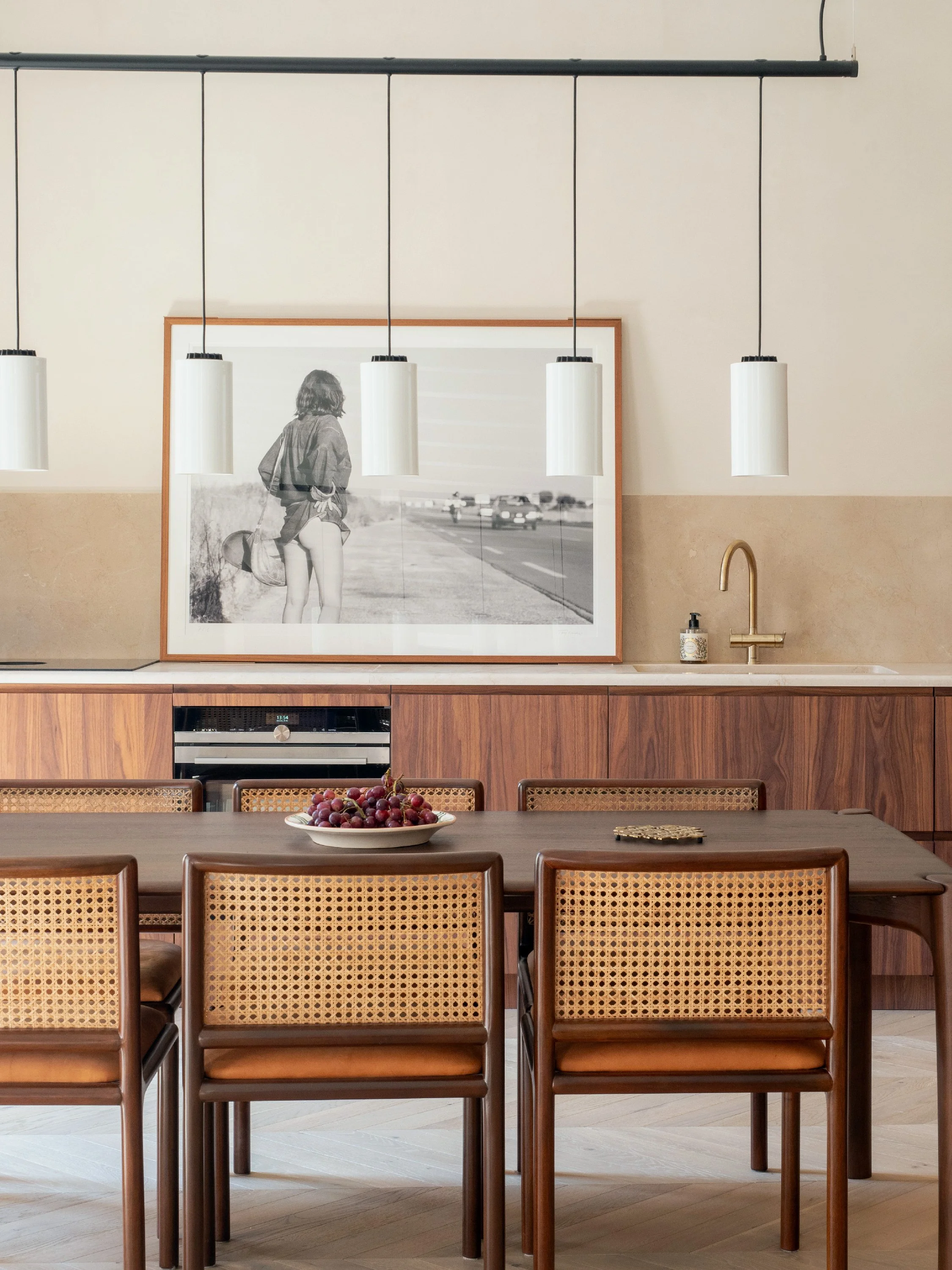 Modern kitchen or dining area with wooden table and chairs, a bowl of grapes, a framed black-and-white photo on the wall, a gold faucet, and hanging white pendant lights.