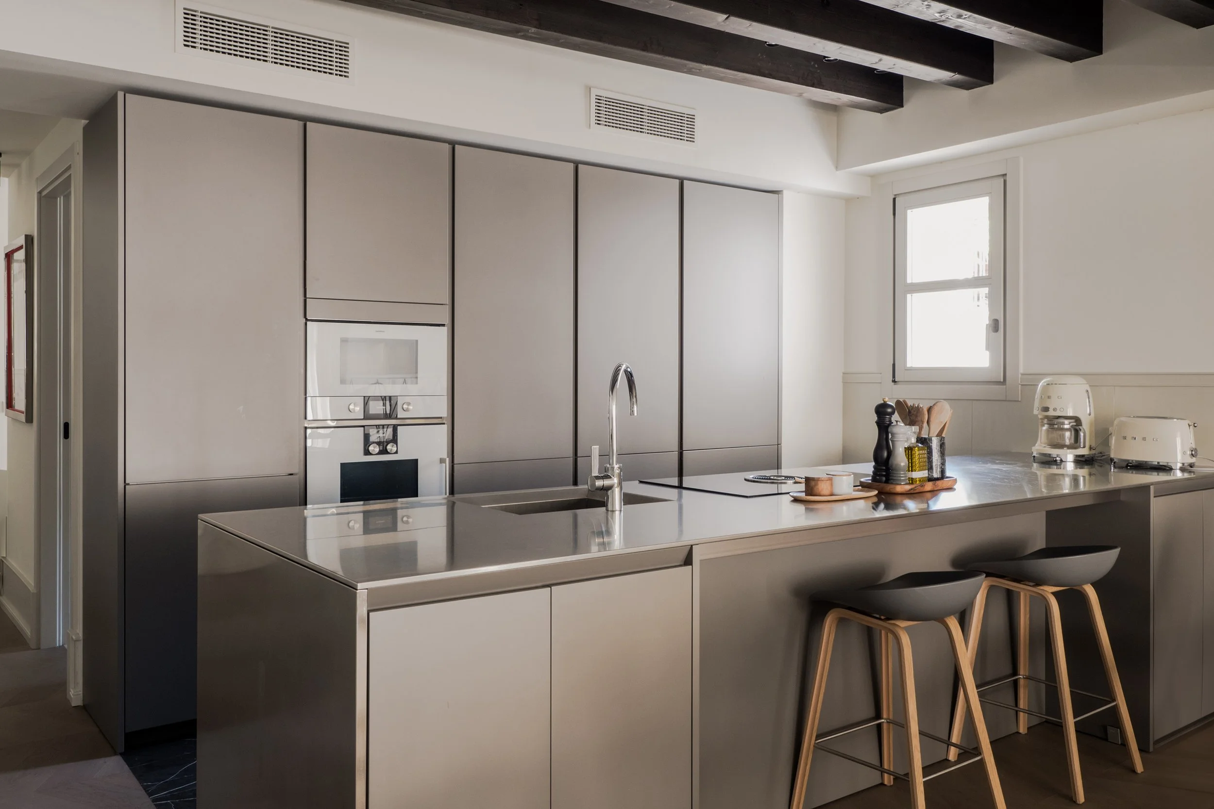 Modern kitchen with gray cabinets, a kitchen island with a sink and faucet, wooden chairs at the island, a window, and kitchen appliances including a stand mixer and pepper mill.