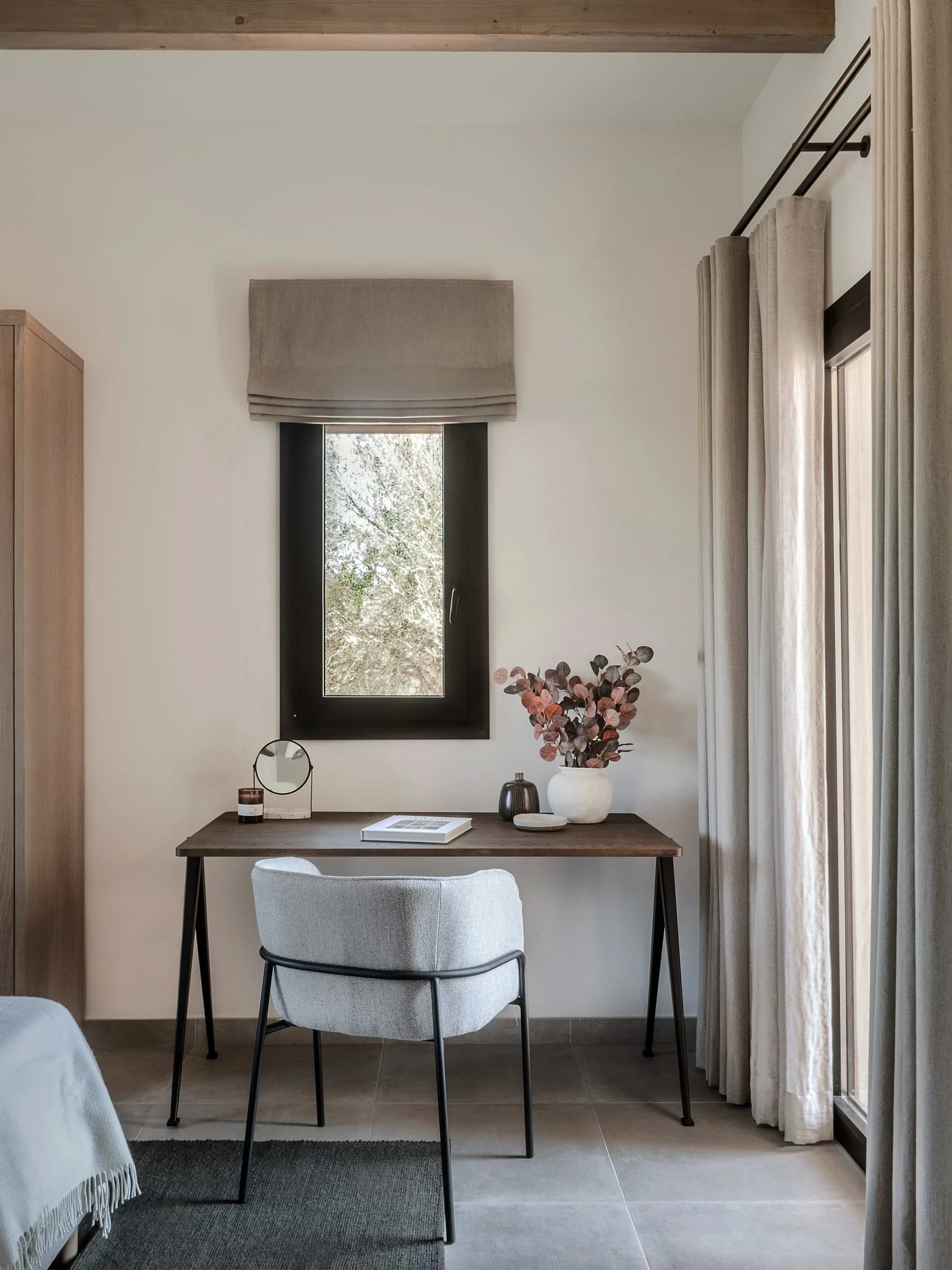 A desk with a white chair, decorated with vases, a small mirror, and a magazine, against a wall with a window and curtains, and a wooden ceiling.