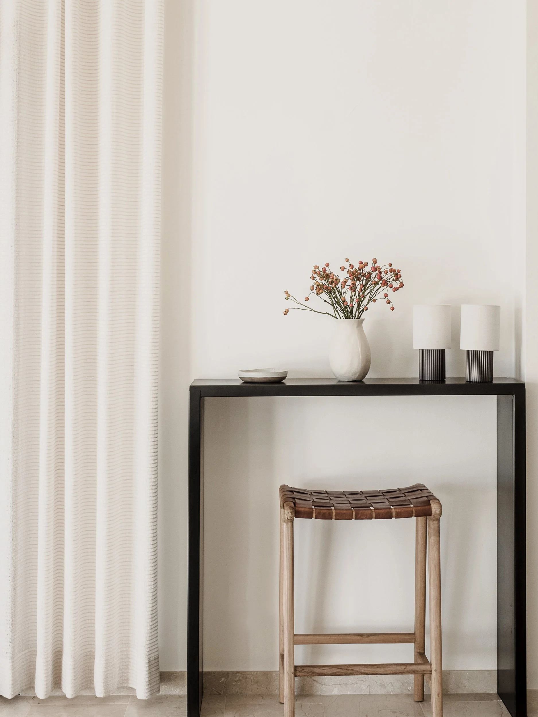 A minimalist interior scene with a black metal table, a white ceramic vase with pink and red dried berries, two white lamps with striped black bases, a light brown woven stool with wooden frame, cream-colored curtains on the left, and a plain off-whi