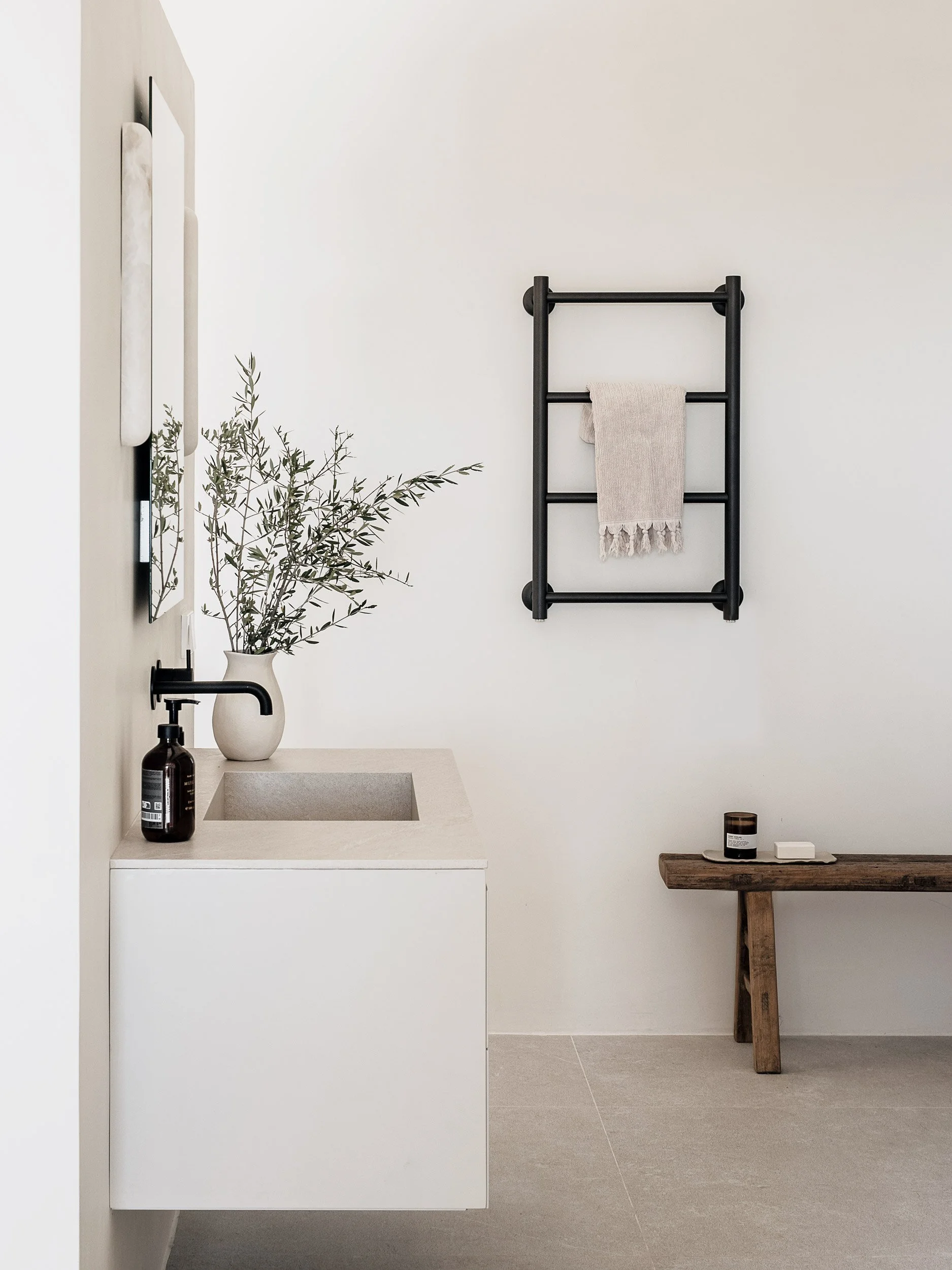 Minimalist bathroom with white walls, a white sink, a black towel rack with a beige towel, a small wooden table with a candle and soap, and a potted plant on the sink counter.