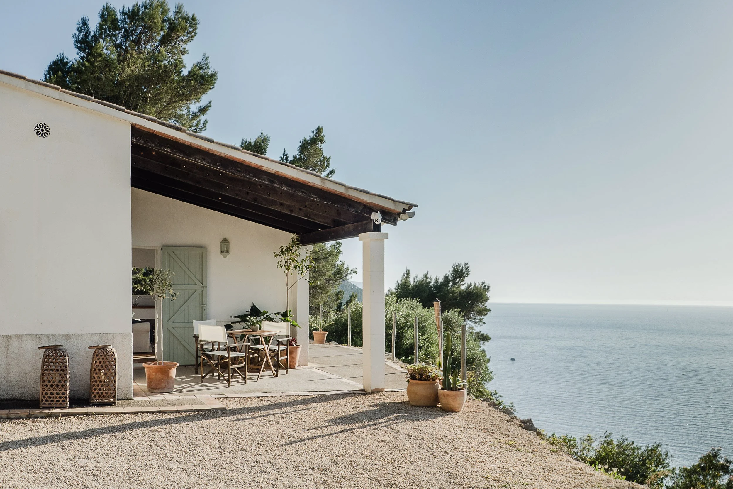 A white house with a patio overlooking a body of water and trees, decorated with potted plants and outdoor furniture under a clear sky.