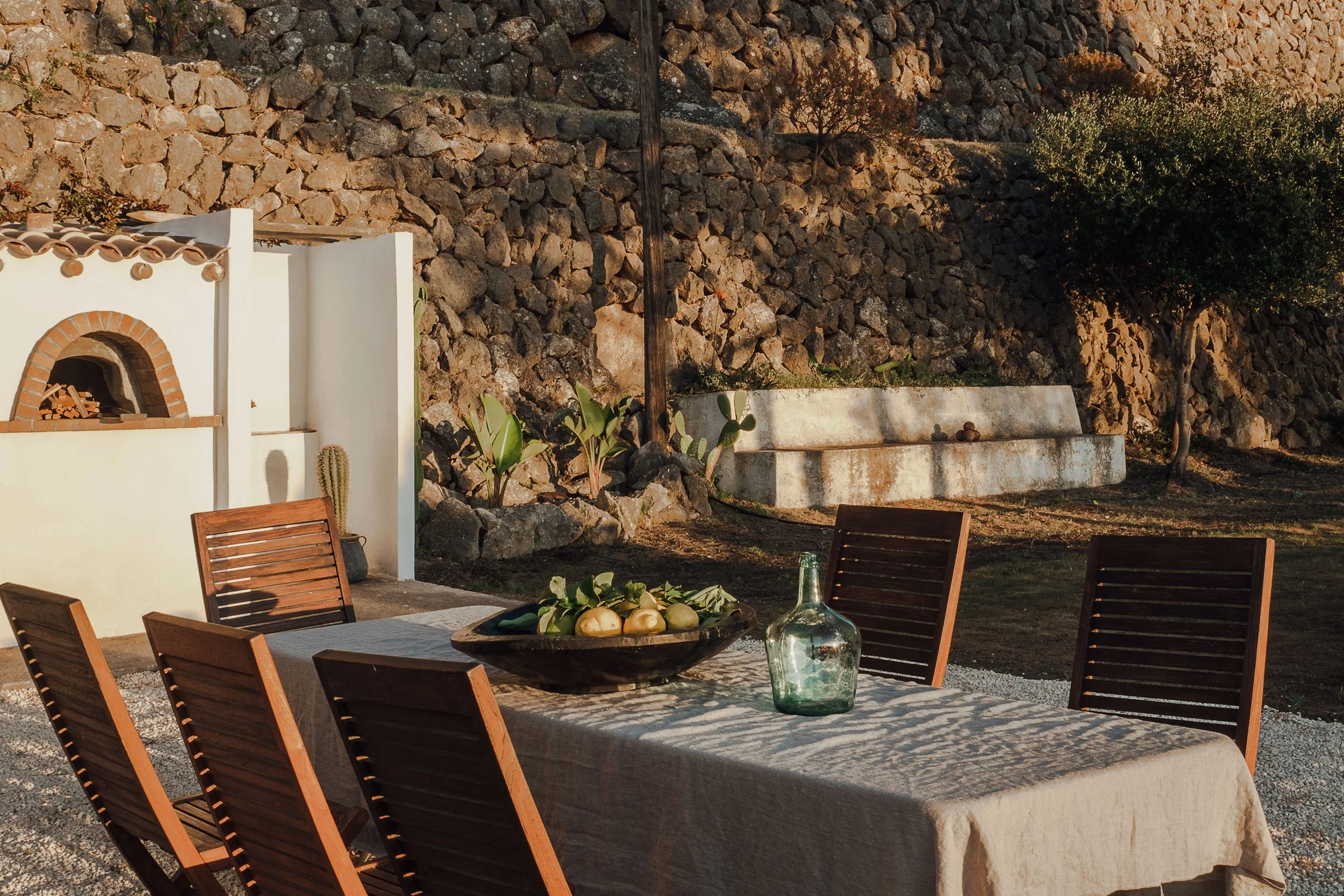 Outdoor dining area with wooden chairs around a table, a bowl of fruits, and a glass bottle, set against a stone wall and trees in the background, lit by warm sunlight.