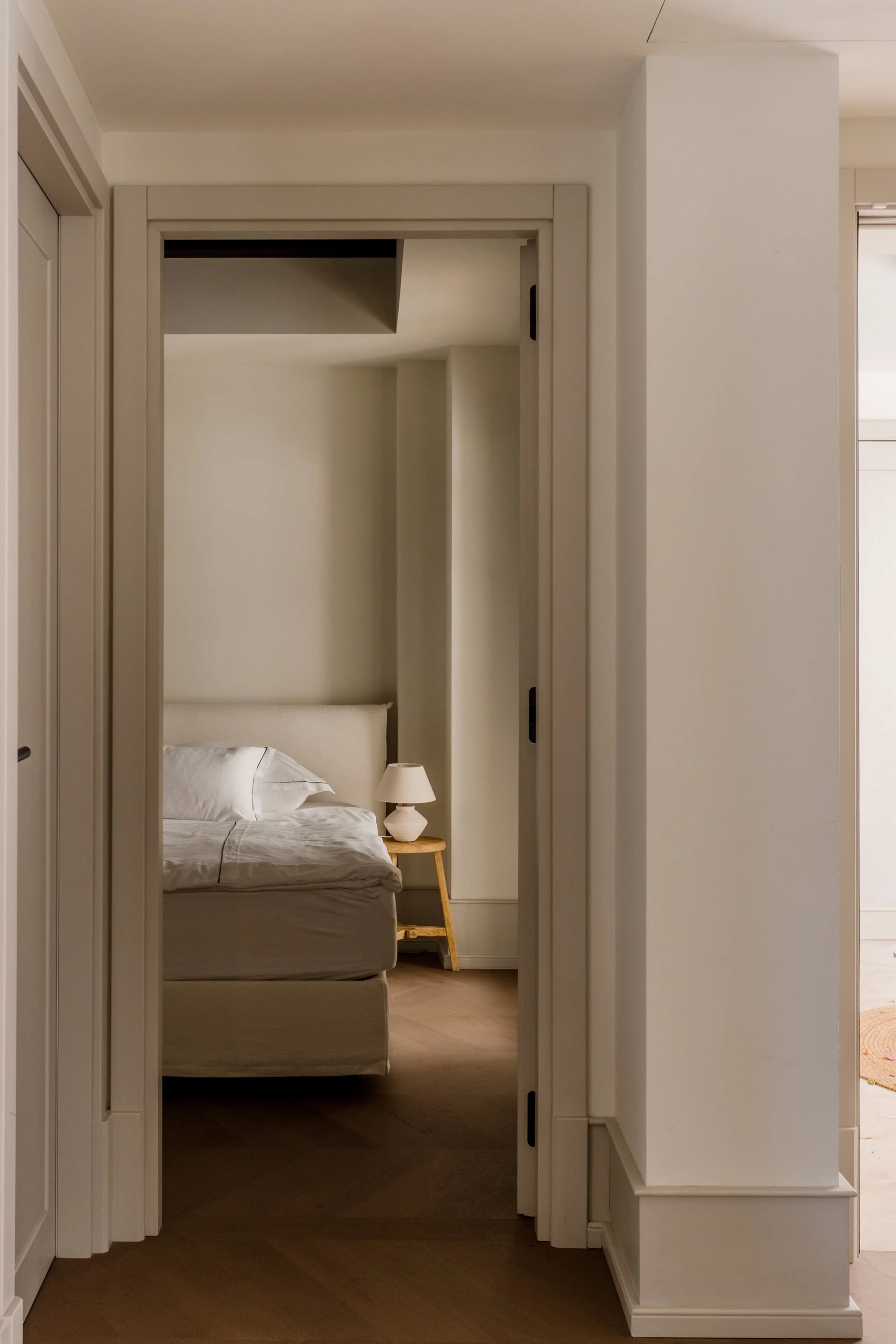 A view of a bedroom through a doorway, showing a bed with white bedding, a bedside table with a white lamp, and beige walls and headboard.