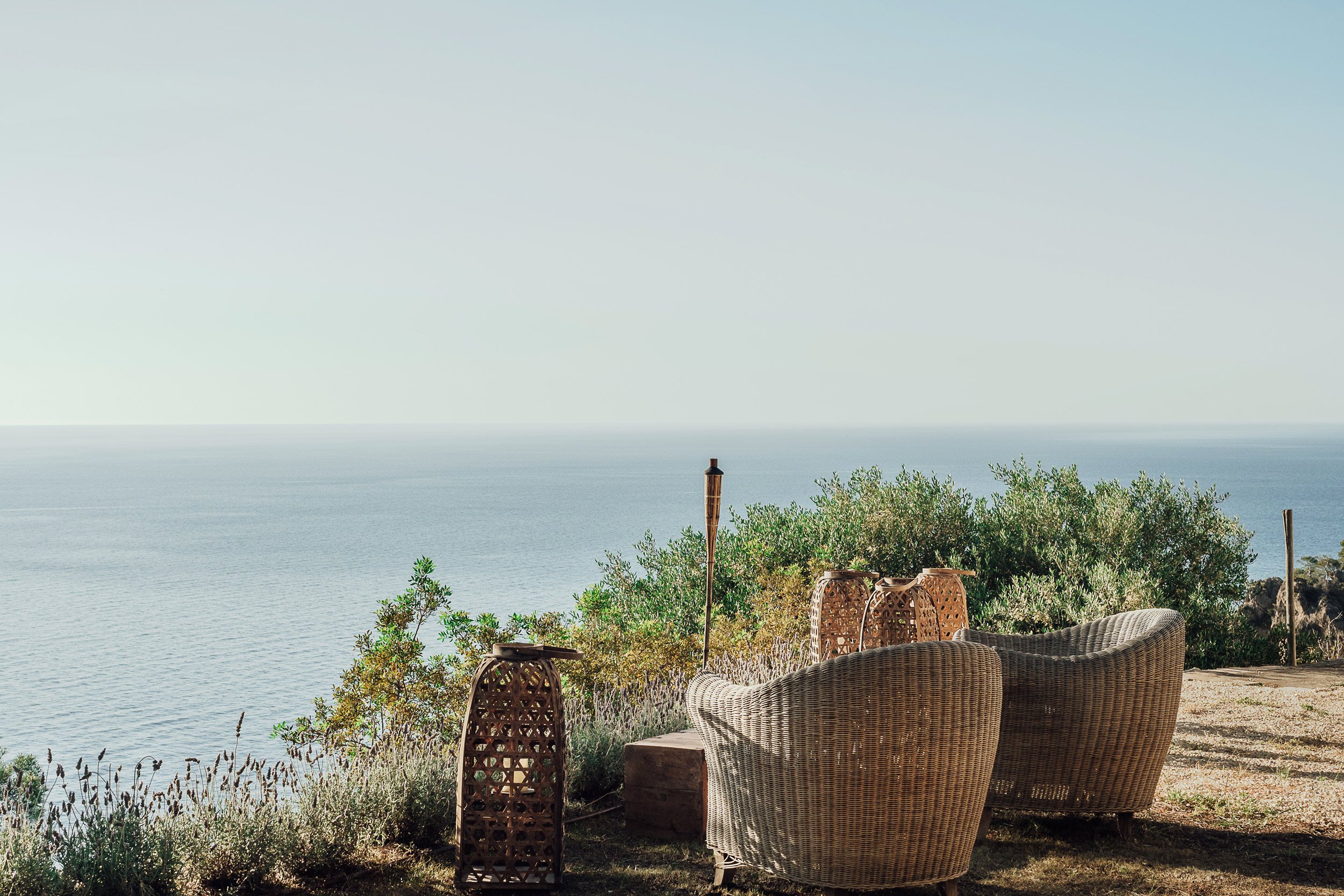 A serene overlook with wicker chairs and decorative lanterns facing the ocean, greenery in the foreground, under a clear light blue sky.