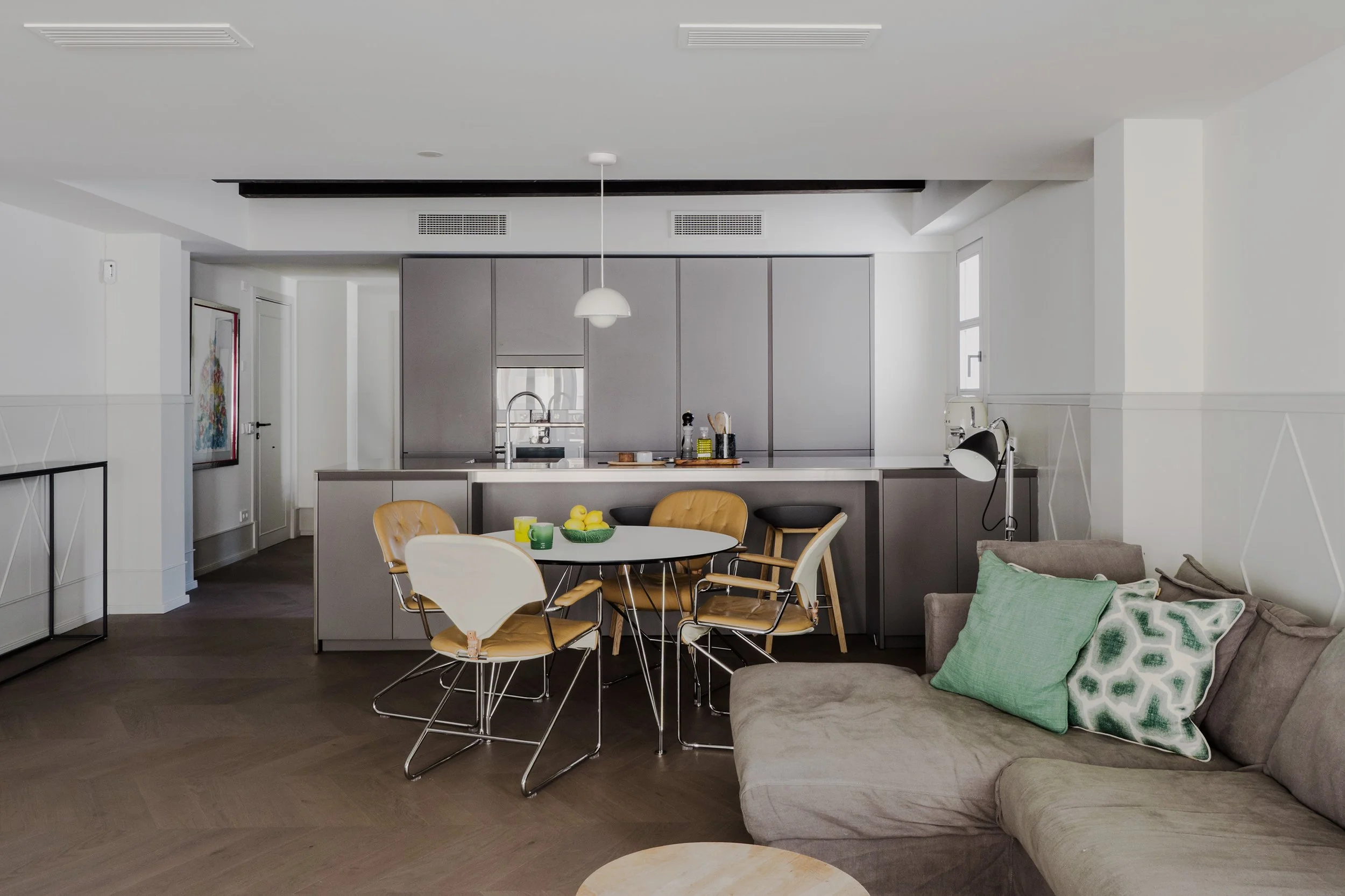 Modern kitchen and dining area with a round table, yellow and white chairs, a gray couch with green pillows, and a white pendant light hanging from the ceiling.