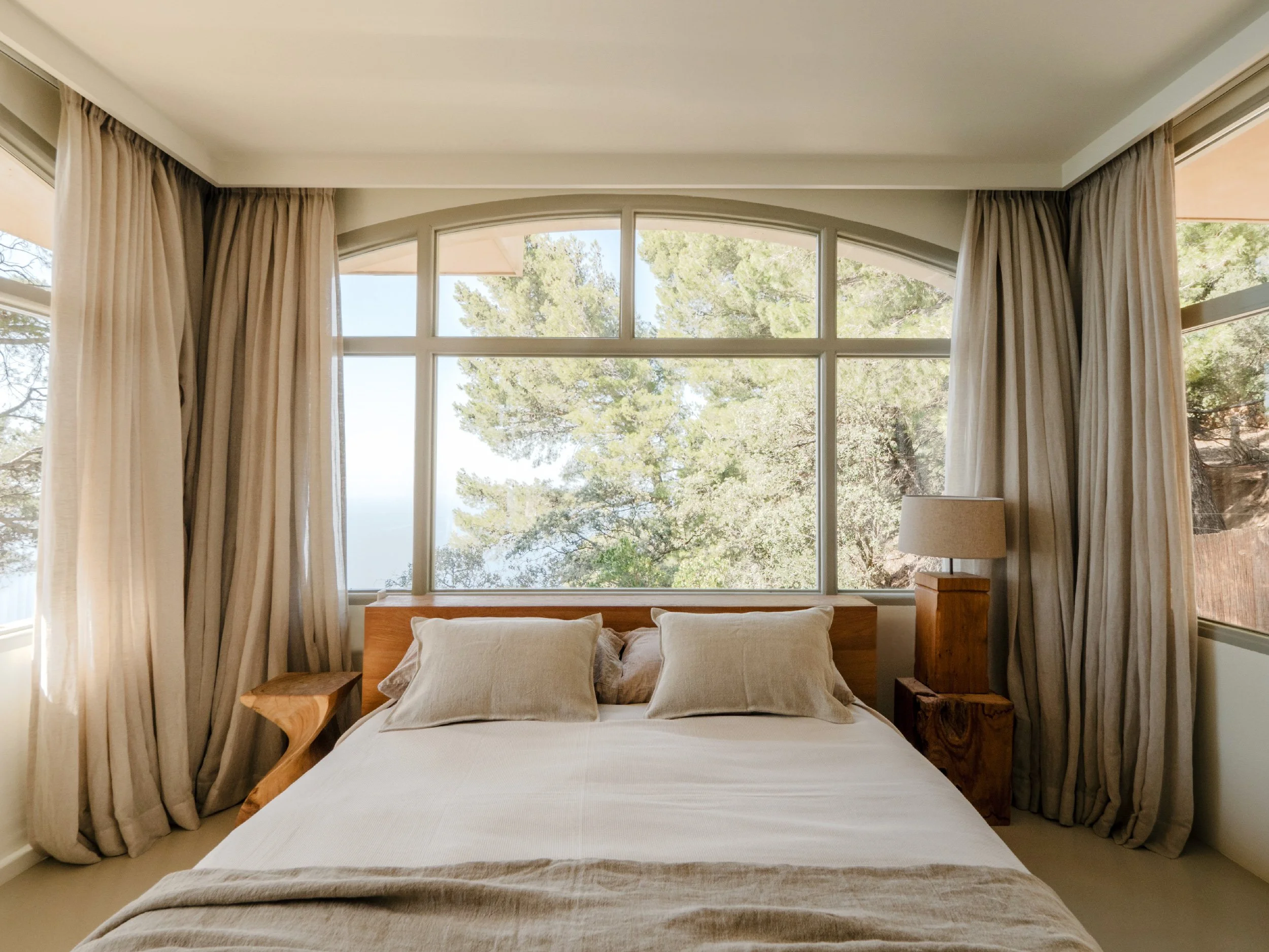 A bedroom with a large bed, beige pillows, and cream-colored bed linens, flanked by wooden side tables, one with a lamp, and large windows with beige curtains revealing trees outside.