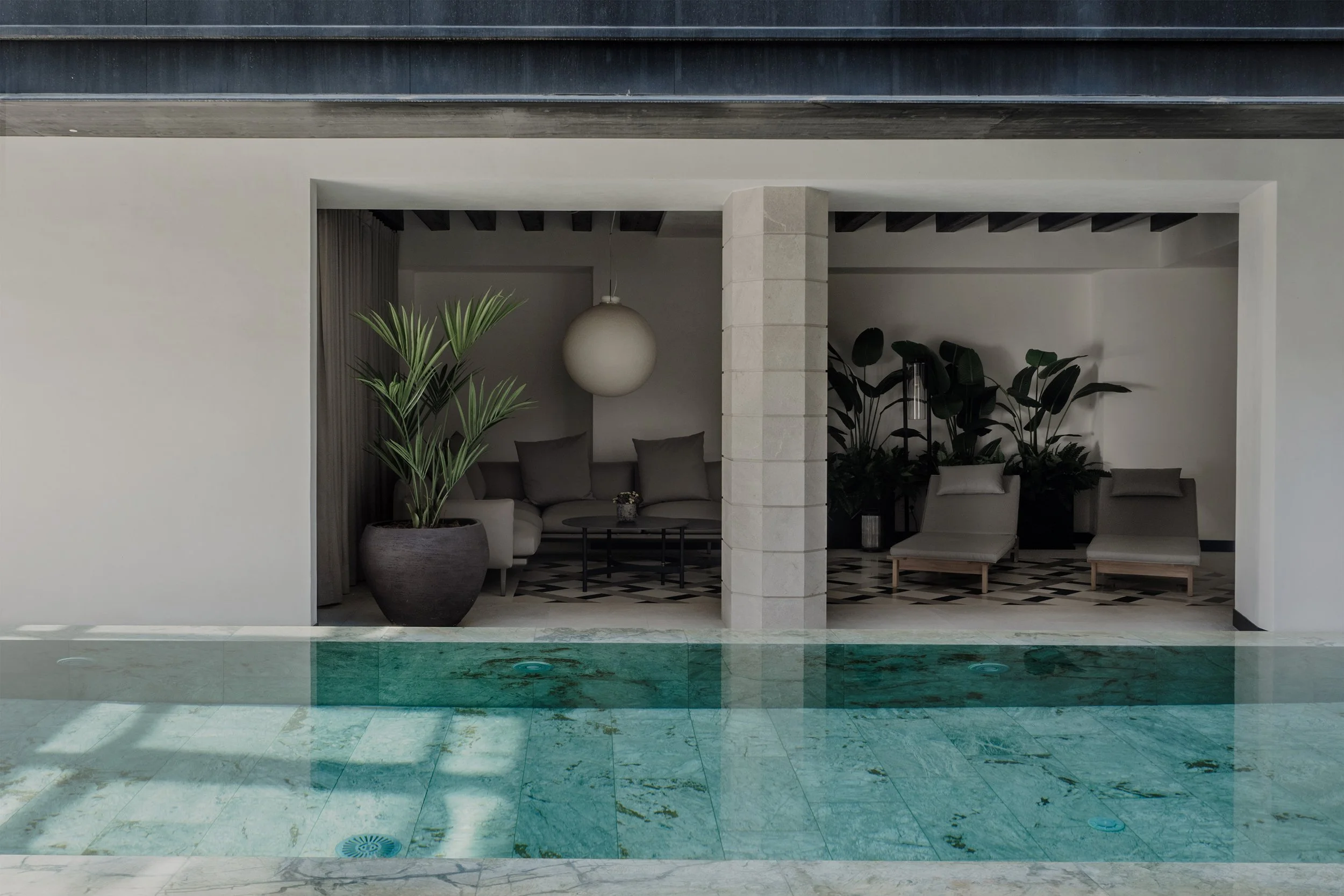 Interior view of a modern space with a small pool in the foreground, decorated with green marble tiles, and a sitting area with beige furniture, large potted plants, and a spherical hanging light fixture.