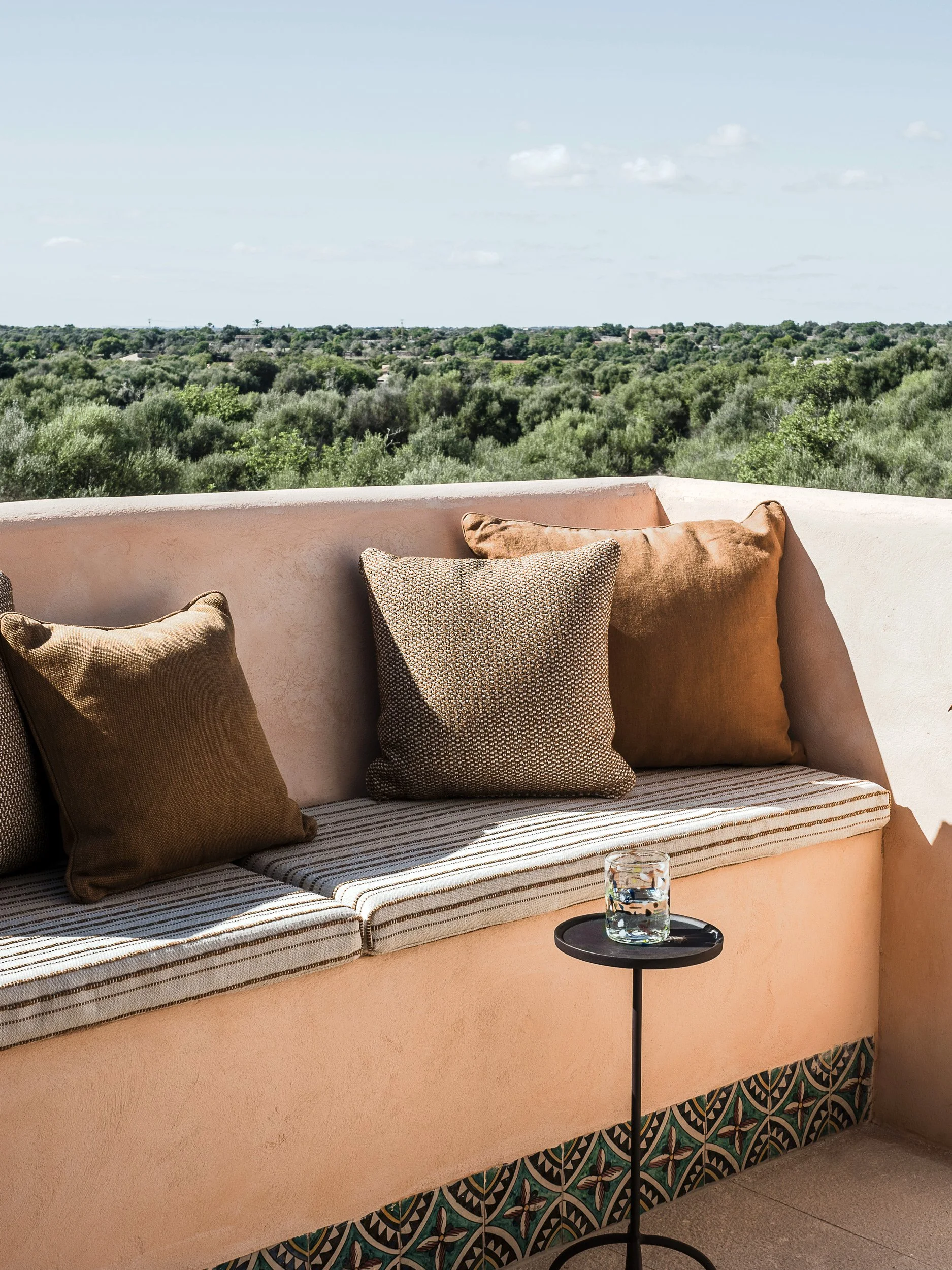 A seating area with beige cushions on a bench, a small black side table with a glass of water, and a view of green trees and a blue sky in the background.