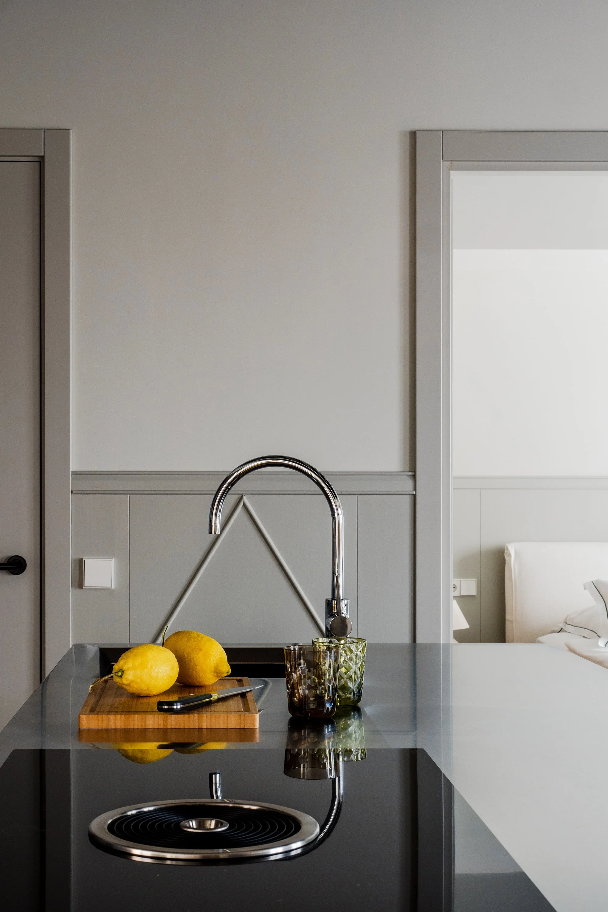 Modern kitchen with a black countertop and sink. On the countertop, there are three yellow lemons on a wooden cutting board, a knife, a glass with a geometric pattern, and a glass with a decorative stem. The background shows a wall with a doorway and