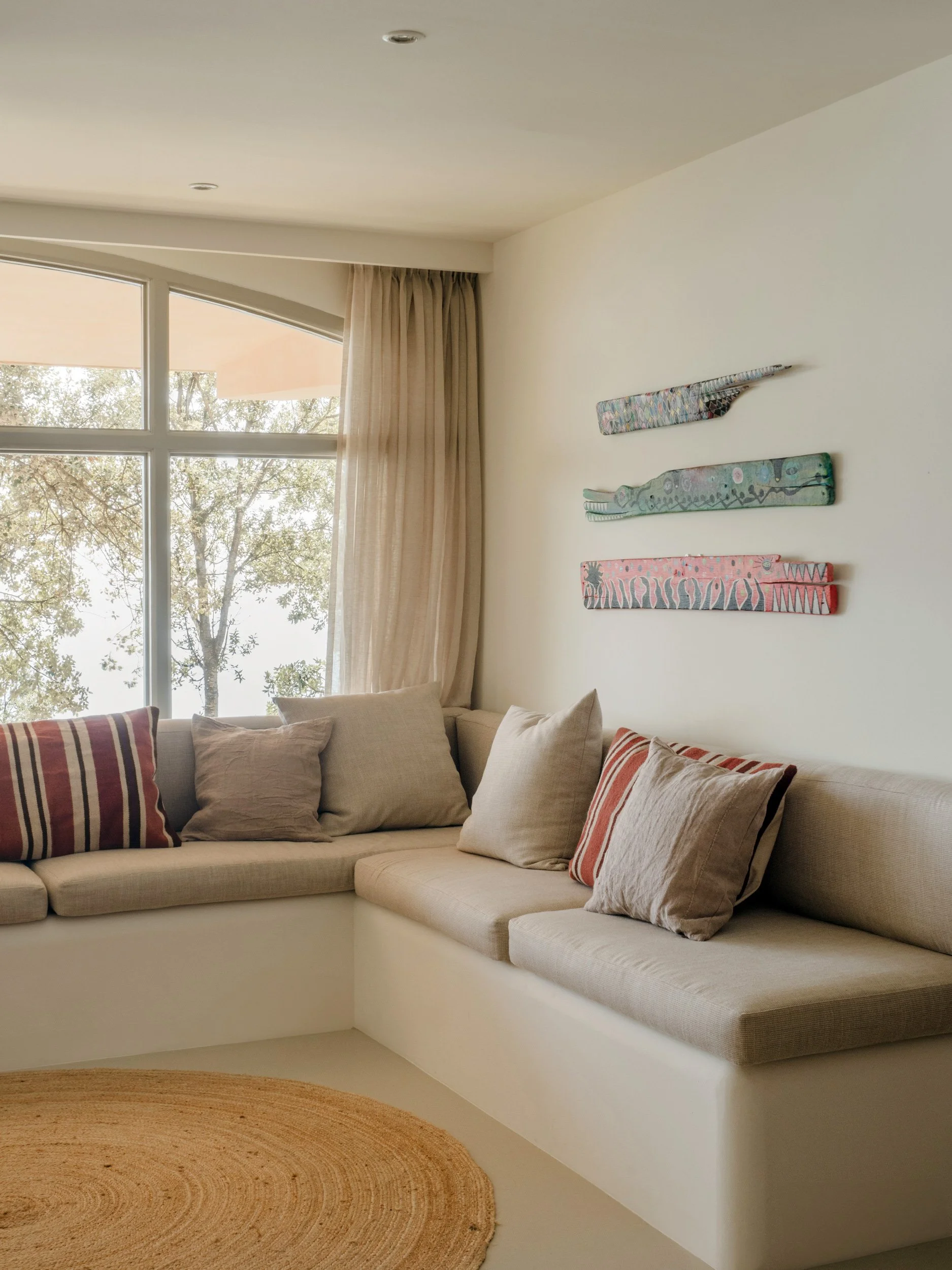 A beige built-in corner sofa with multiple throw pillows in front of a large window with beige curtains and a view of trees outside. A round woven rug is on the floor.