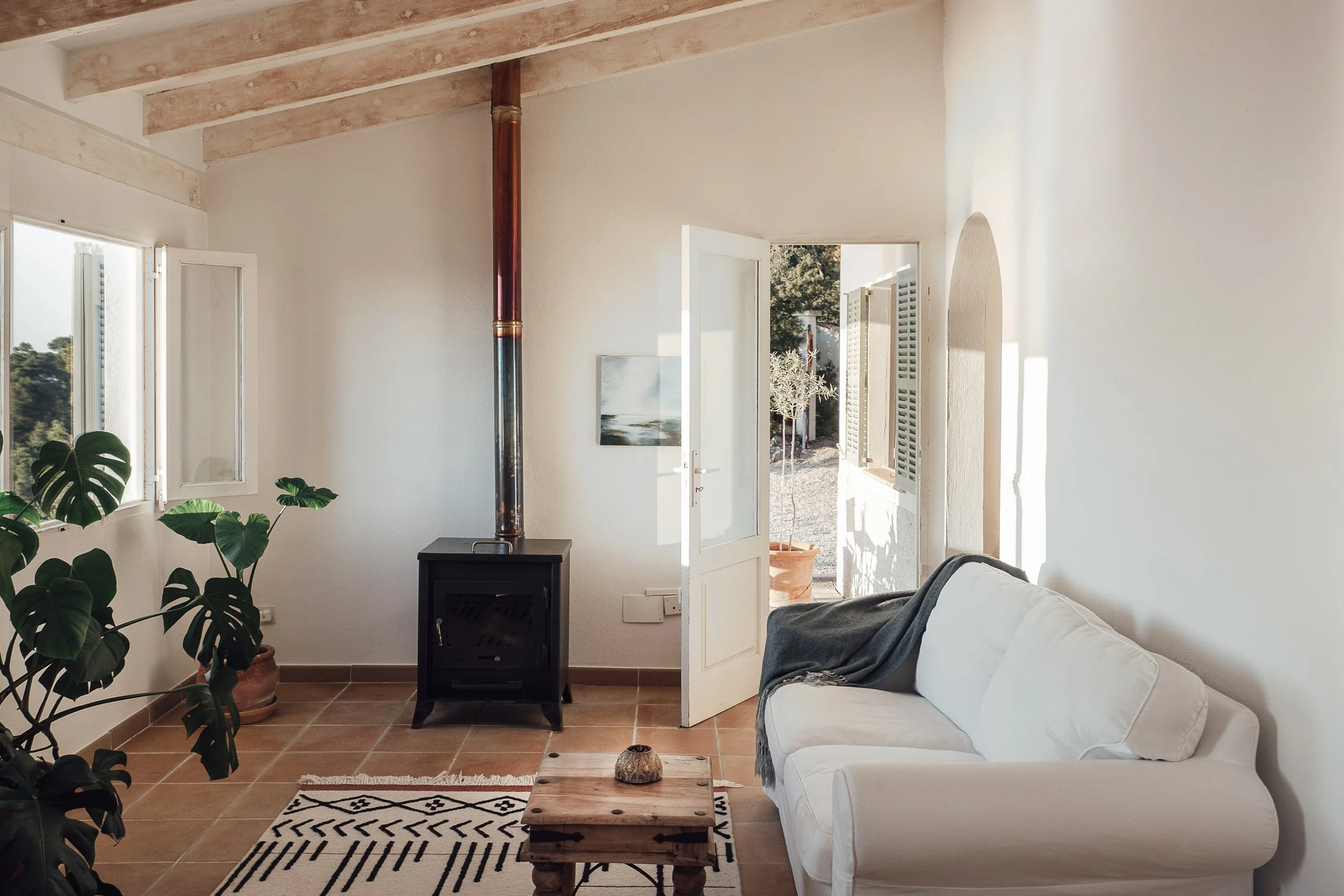 Living room with white walls, a white sofa with a blanket, potted plants, a wooden coffee table, a small black stove, and an open door leading outside. Sunlight streams through the windows, casting shadows.