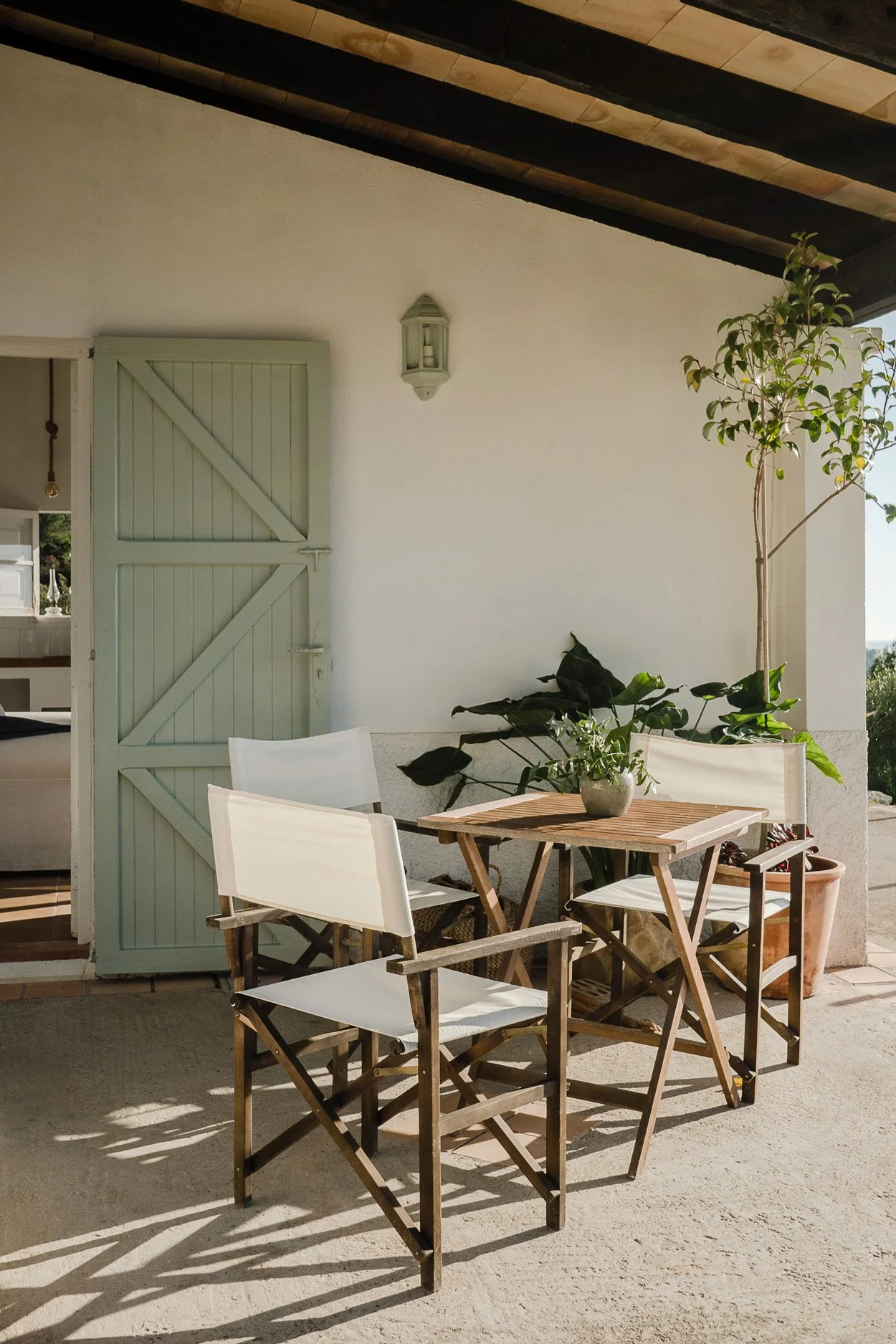An outdoor patio with a wooden table, three white chairs, potted plants, and a light green door, under a sloped ceiling with wooden panels and a wall-mounted lantern.