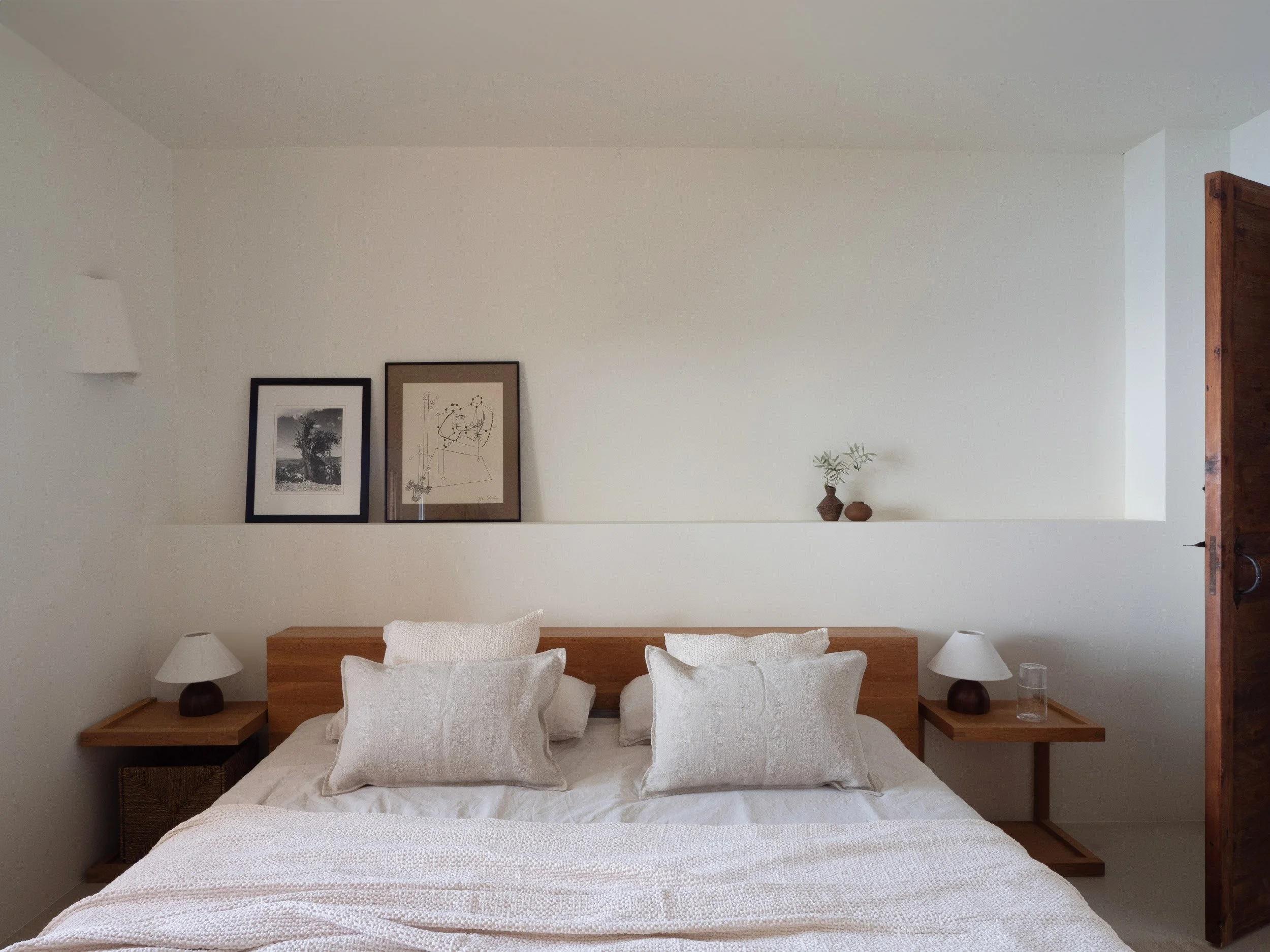 A minimalist bedroom with a bed, two side tables with lamps, three framed pictures on the wall, two vases with plants on the right wall shelf, and a glass on the bedside table.