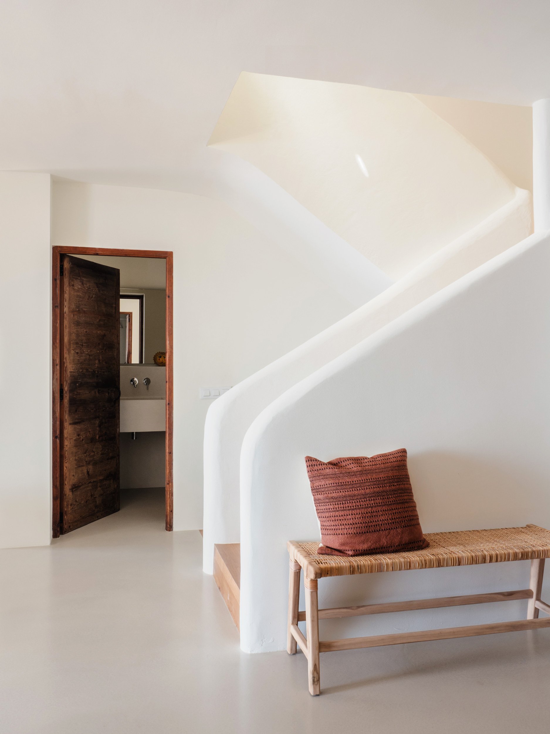 Interior of a minimalist room with white walls and a curved architectural feature, a wooden bench with a woven seat and a rust-colored pillow, visible doorway leading to another room with a sink and a small window.