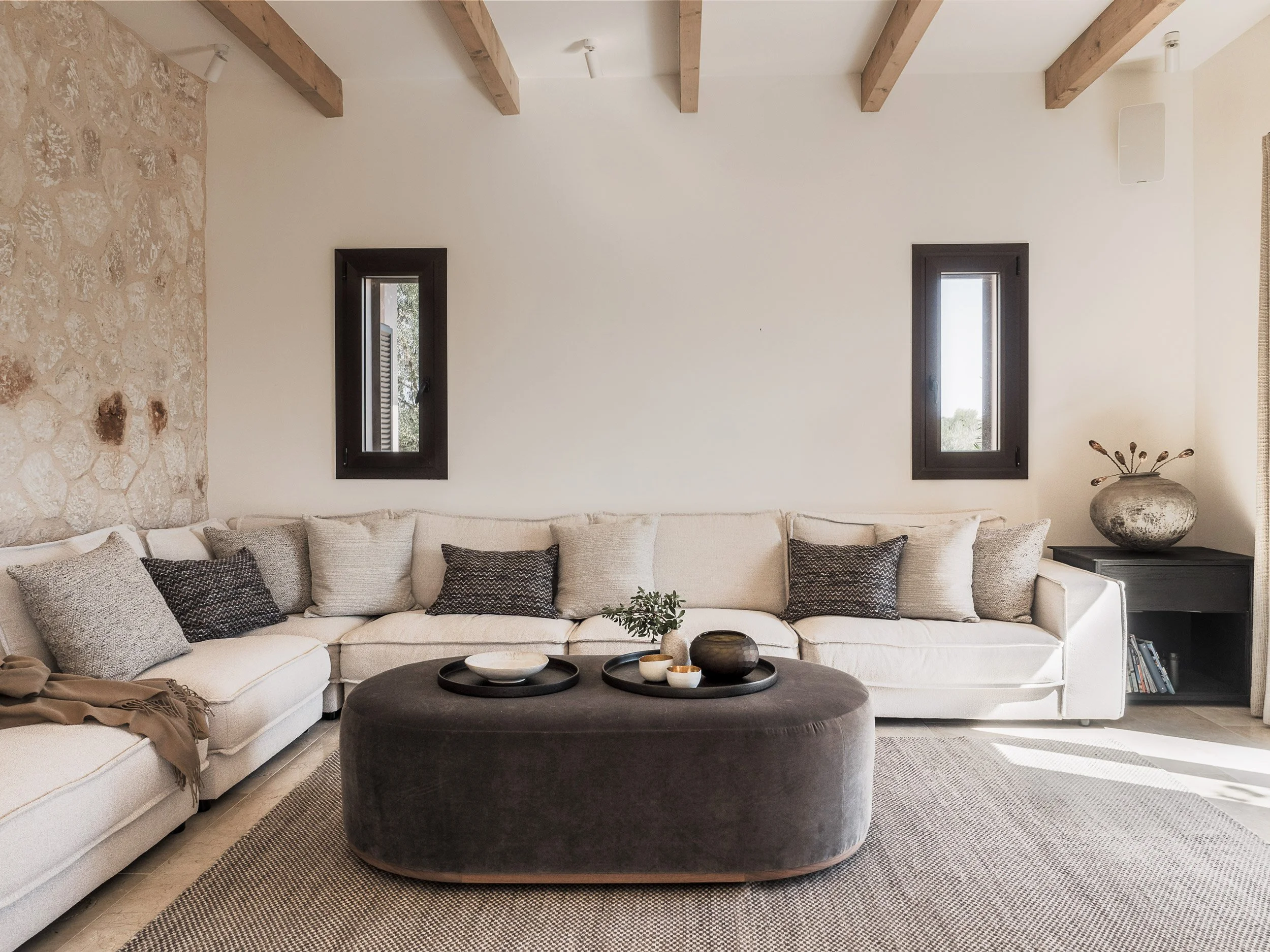 A living room with a beige sectional sofa, pillows, a black round ottoman, a tray with bowls, a stone wall on the left, two small windows, a vase with dried plants, and a shelf with books.