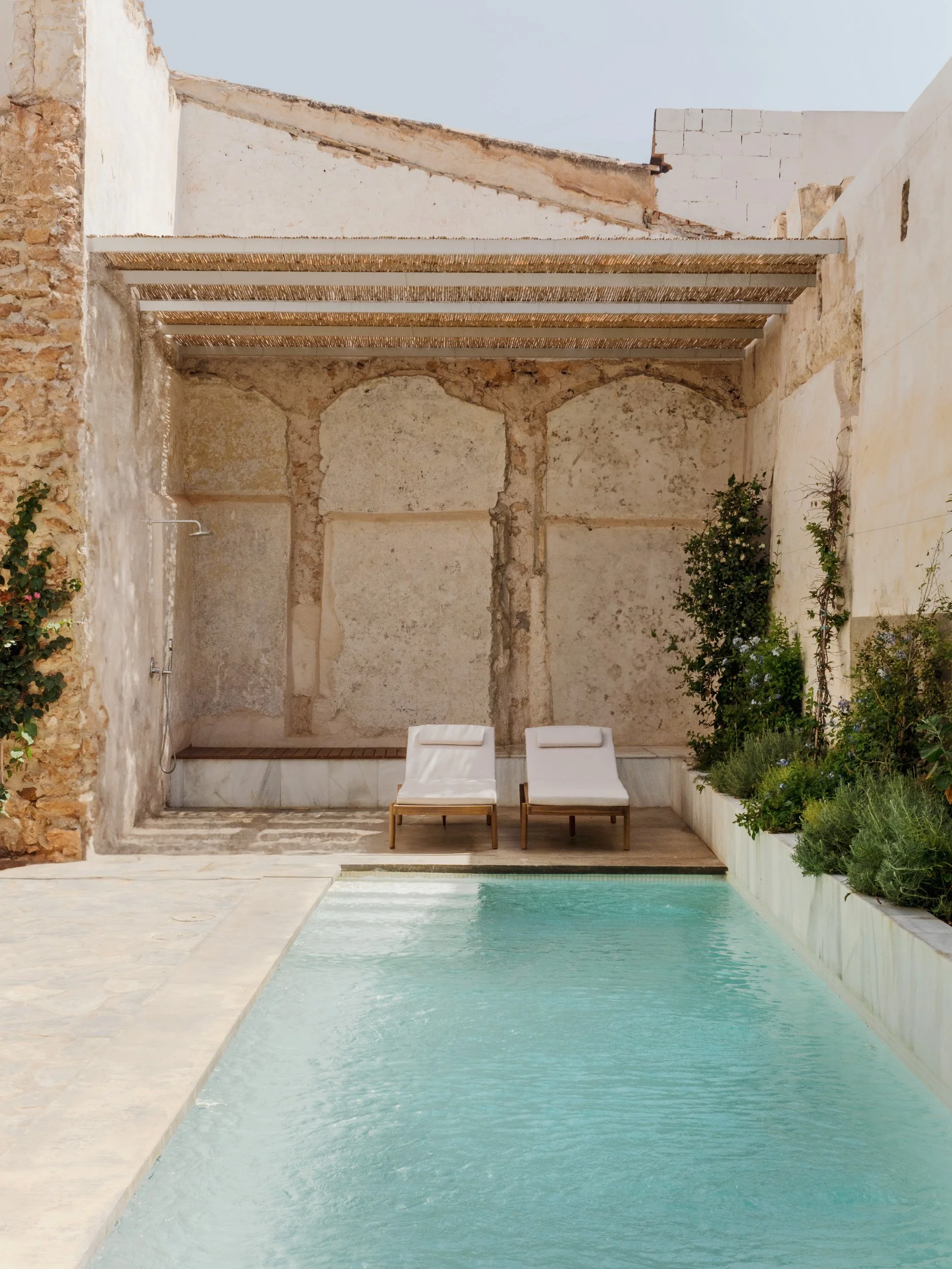 A minimalist outdoor pool area with two white lounge chairs on a wooden deck, surrounded by lush green plants, with an old stone wall and a partly covered canopy in the background.