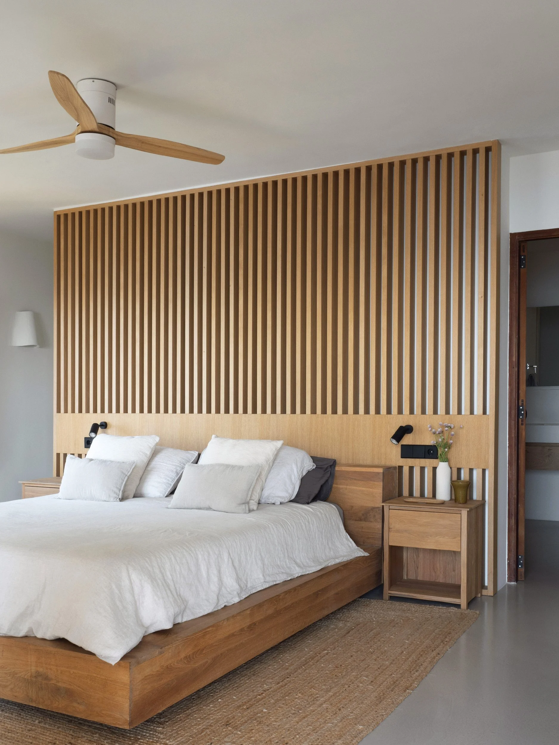 A bedroom with a white bed, wooden headboard, black reading lights, a bedside table with a white vase of flowers, and a wooden slatted wall panel behind the bed, with a ceiling fan overhead.