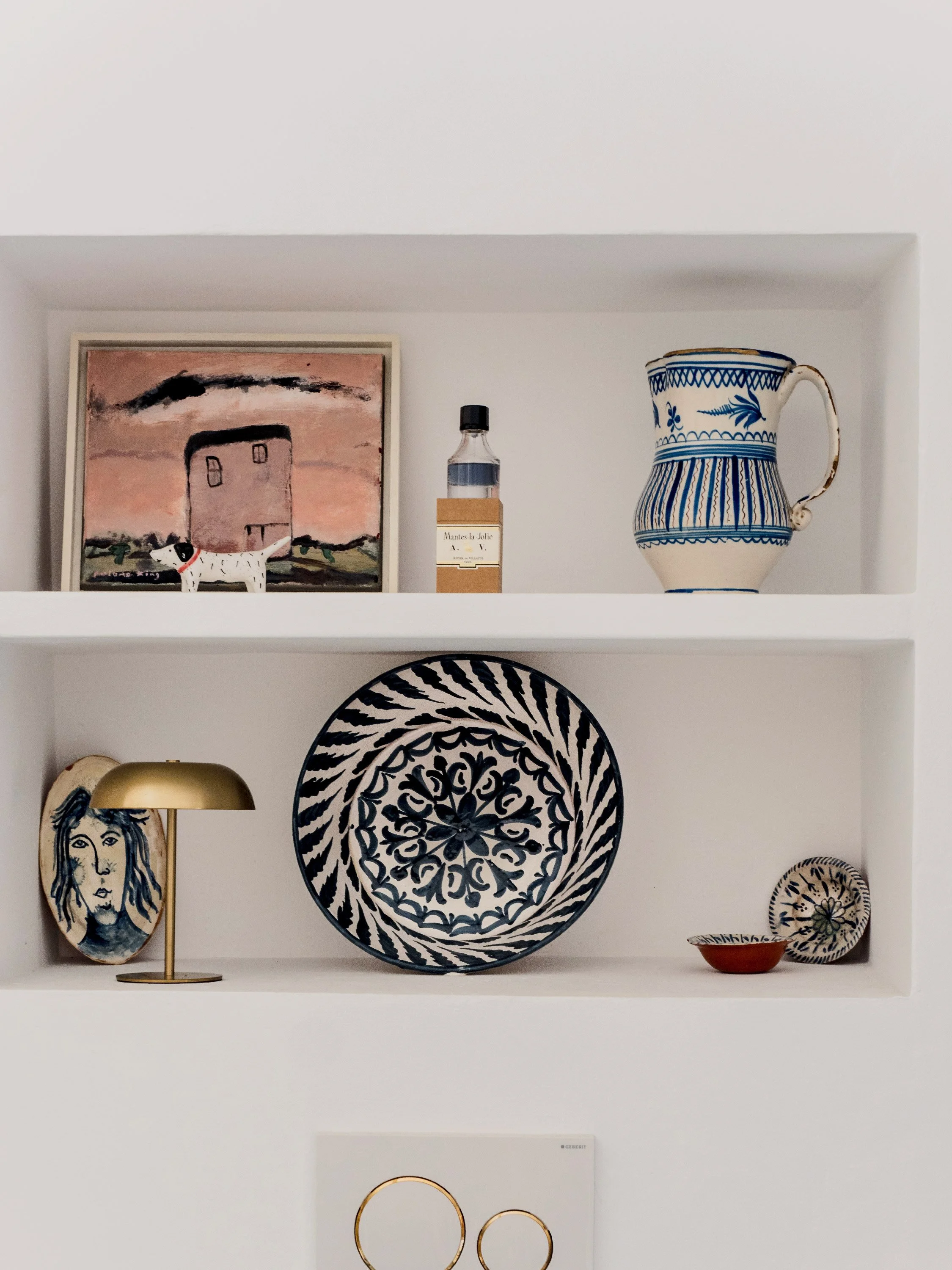 White wall shelf with decorative items, including a painting, a bottle, a blue and white pitcher, a gold lamp, patterned plates and bowls, and a framed art piece.