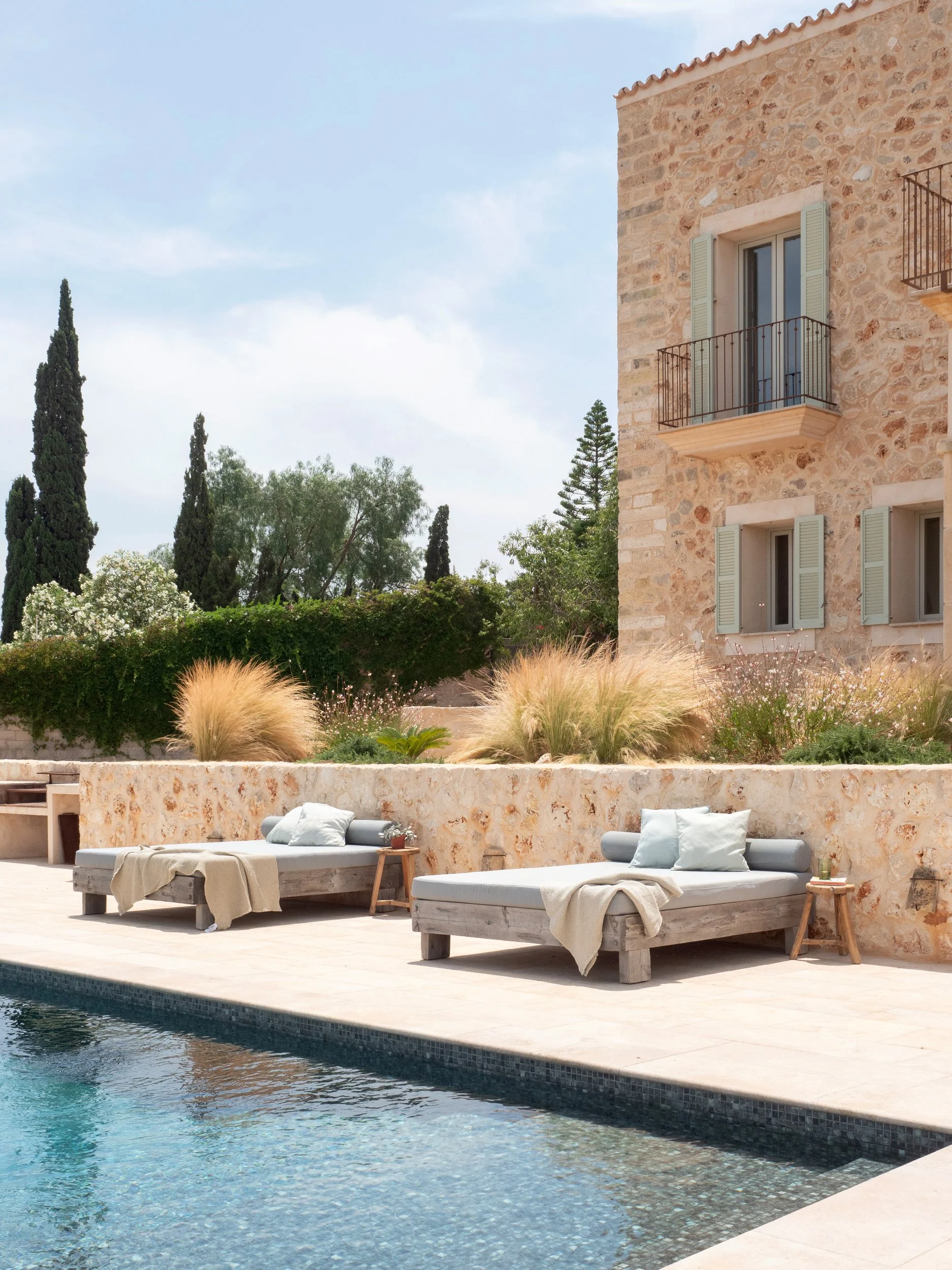 Outdoor scene with a swimming pool, two lounge beds with pillows and blankets, a stone building with small balconies, lush trees, and ornamental grasses under a partly cloudy sky.
