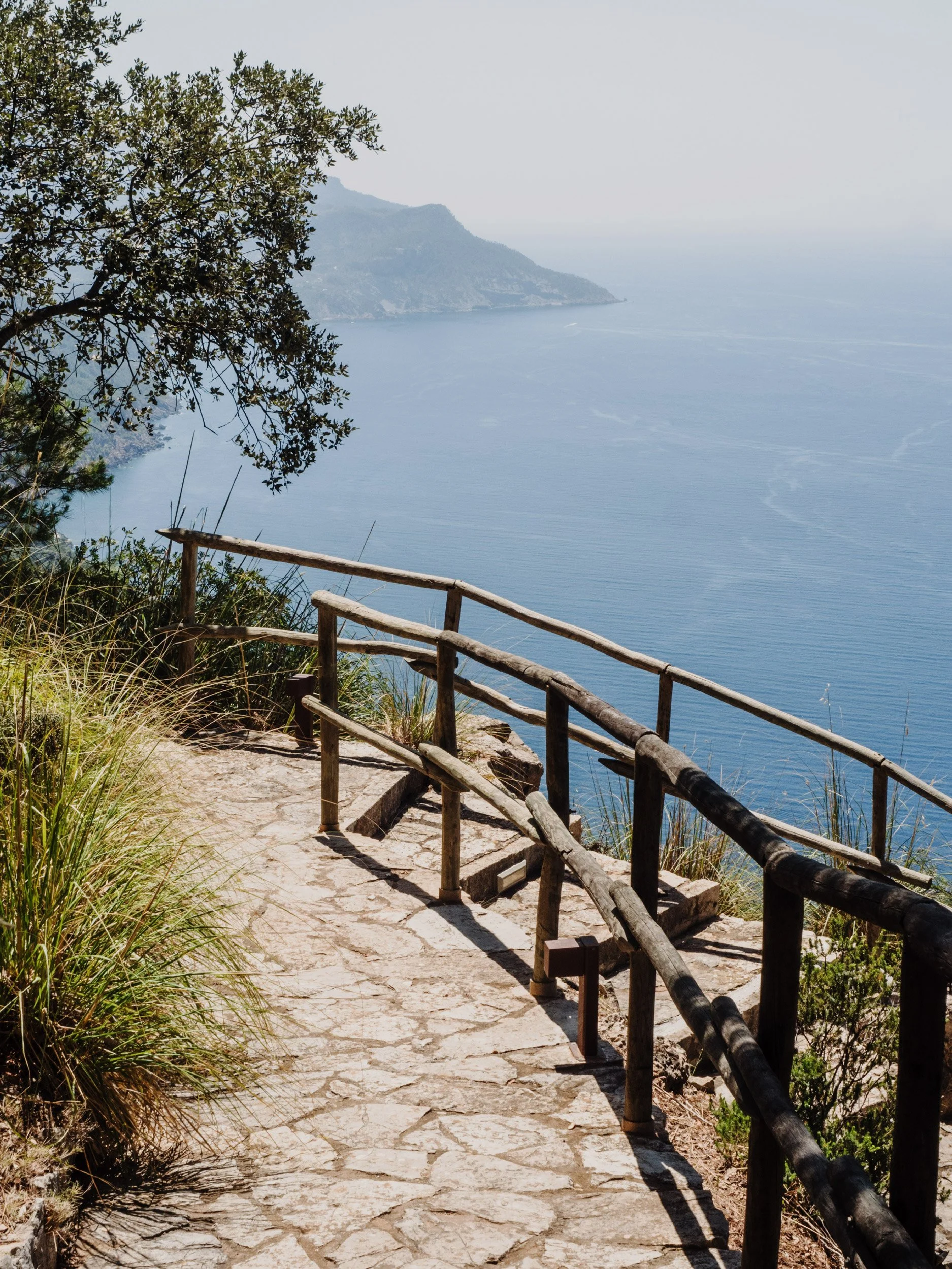 A stone pathway with a wooden railing leads down towards a body of water with hills in the background, trees on the left side, and clear skies.