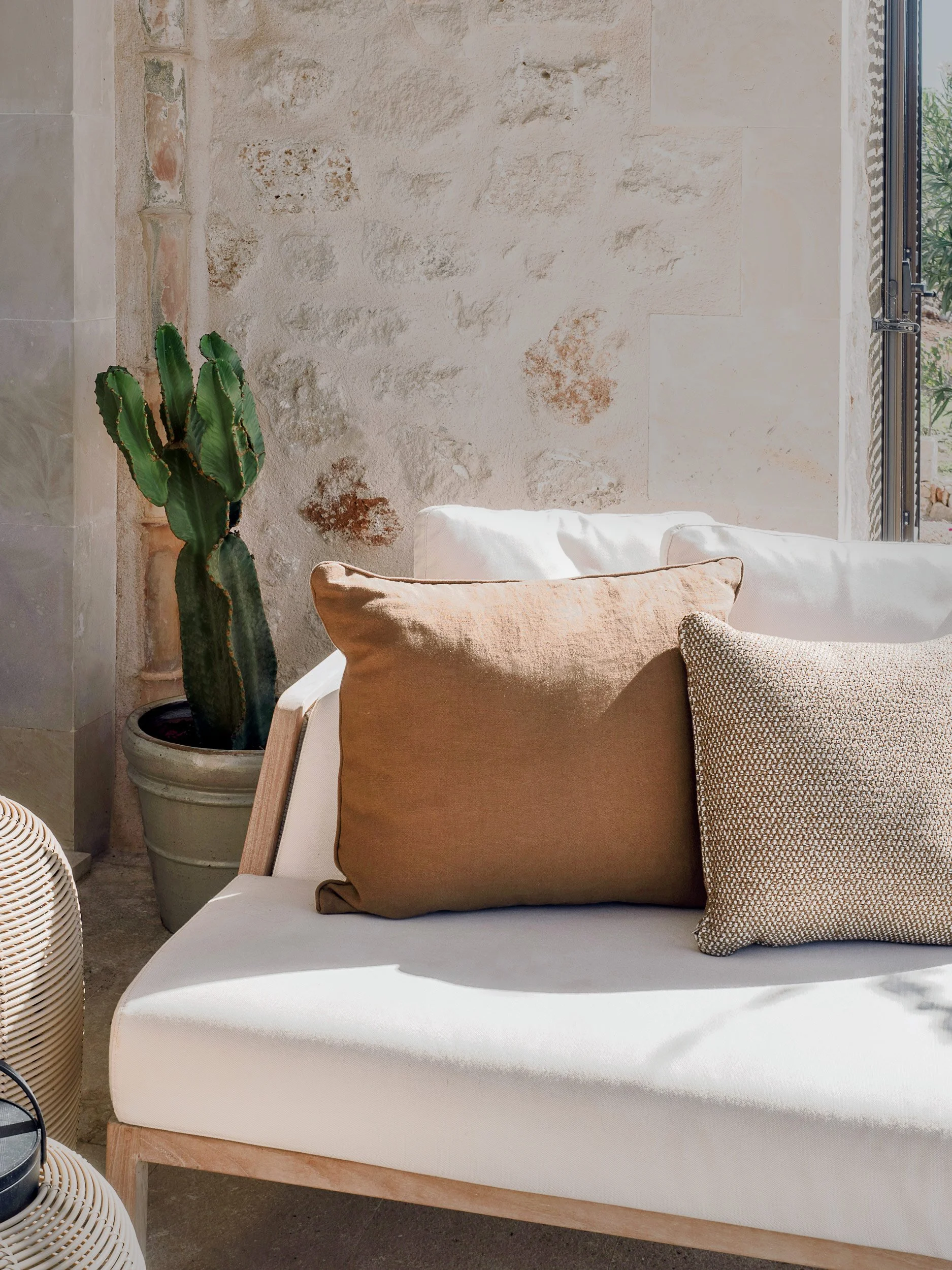 A white outdoor sofa with three pillows in beige, brown, and patterned fabric, positioned next to a cactus plant in a pot, with a stone wall and a window in the background.