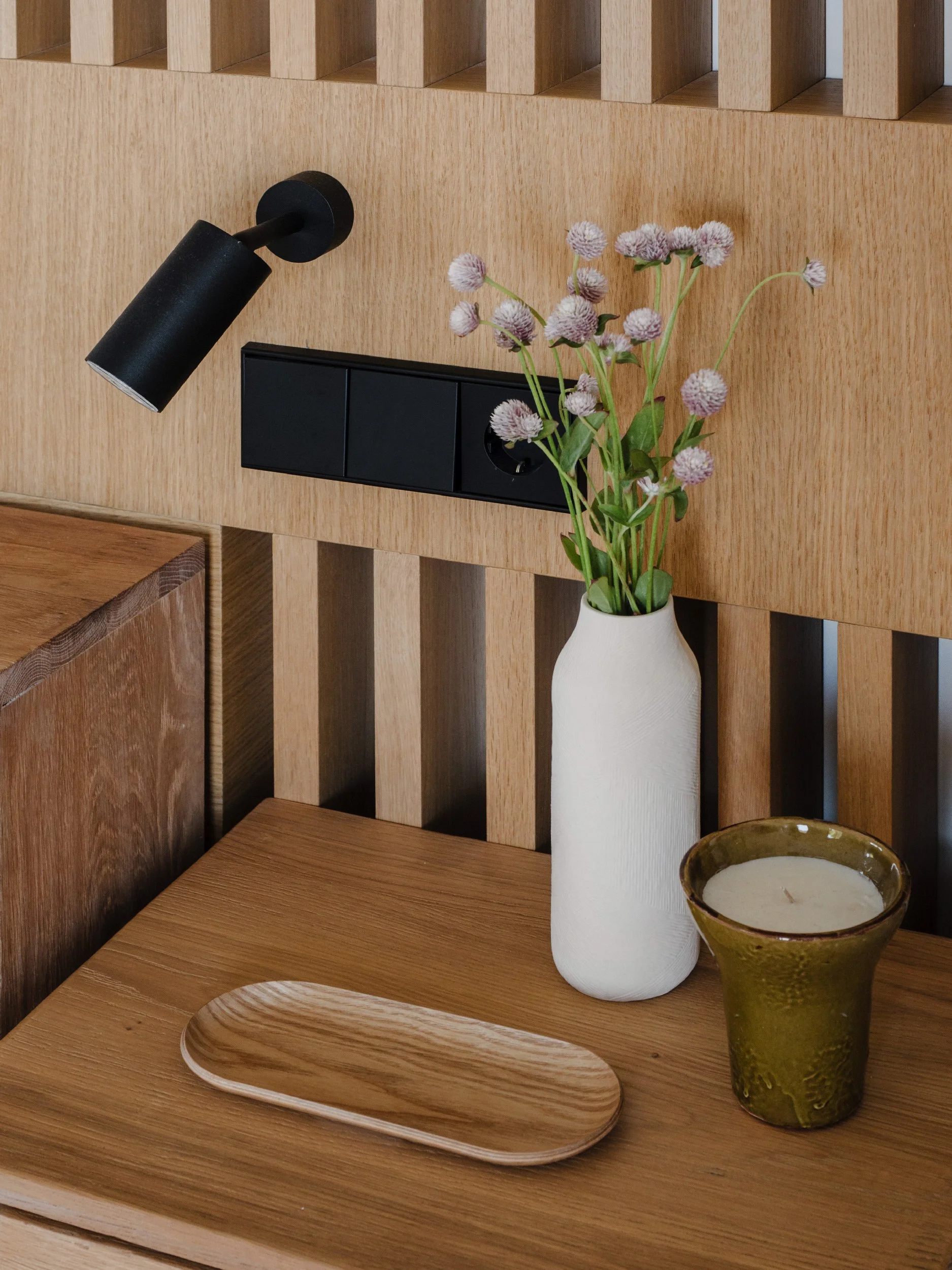 A wooden table with a white vase holding pinkish-purple flowers, a green ceramic cup, and a wooden serving tray.