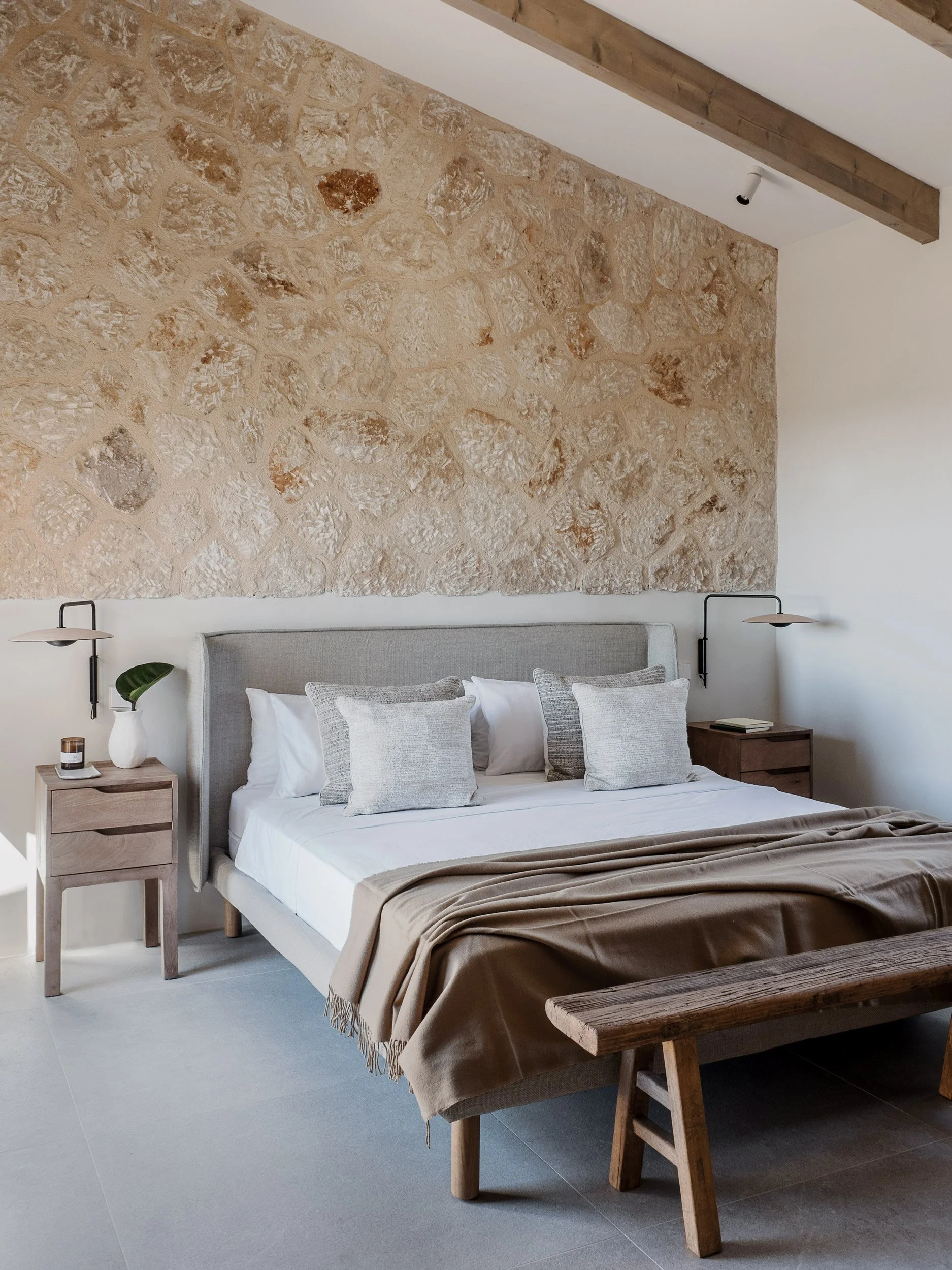 A modern bedroom with a stone accent wall, a bed with white and gray bedding, nightstands with lamps, a potted plant, and a wooden bench at the foot of the bed.