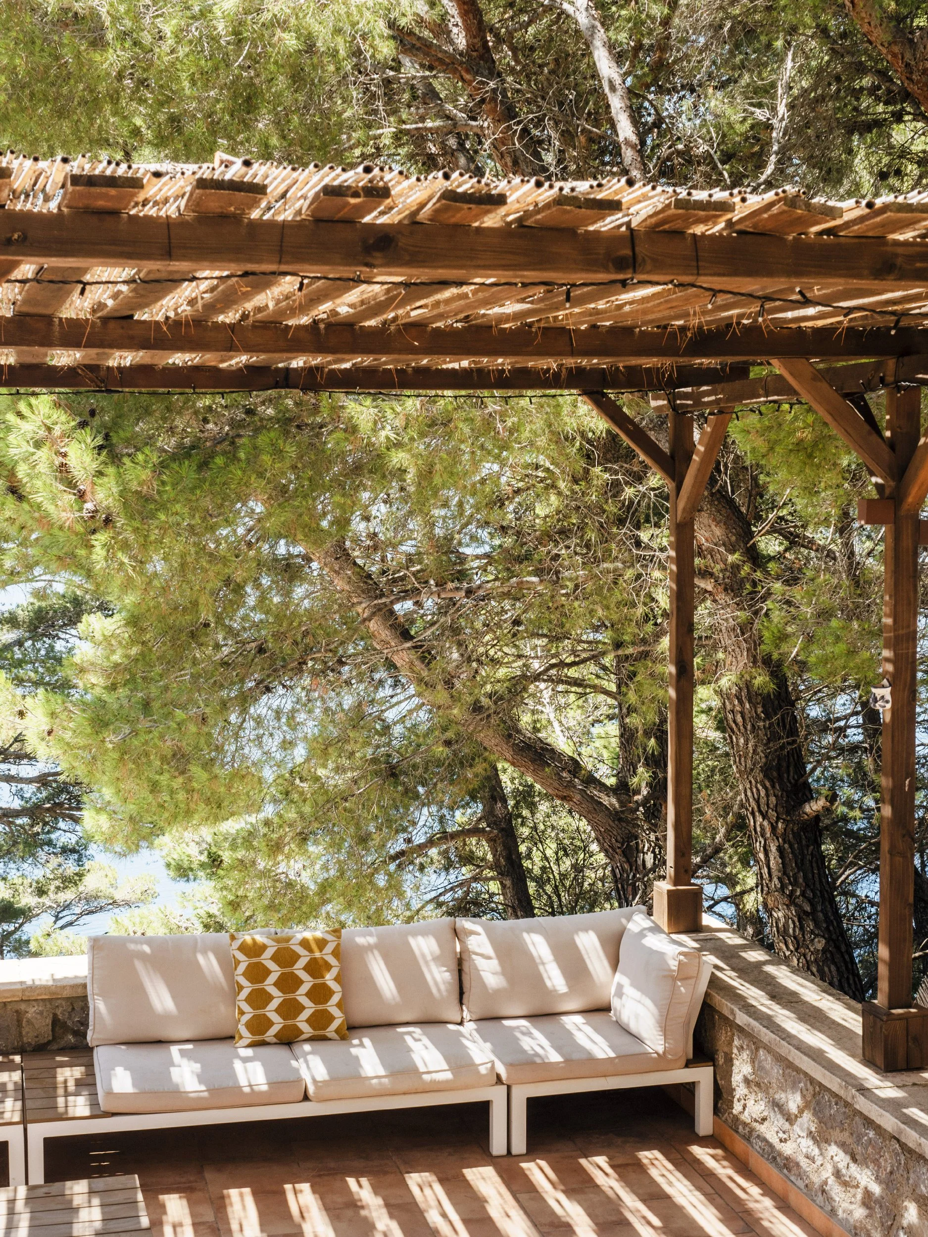 An outdoor patio with a beige cushion sofa, a decorative pillow with a gold honeycomb pattern, and a wooden pergola casting shadows, surrounded by tall green trees.