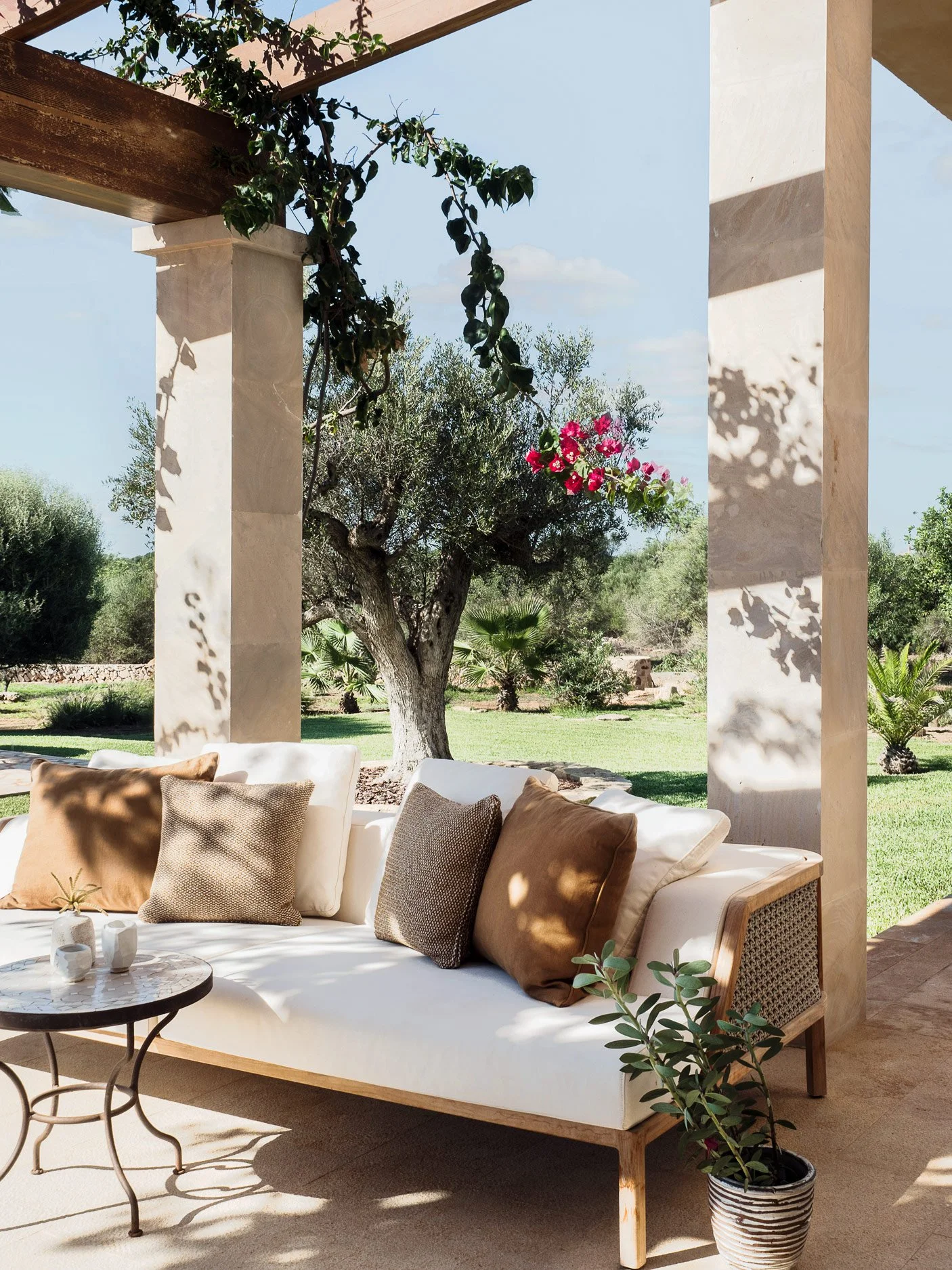 Outdoor patio area with a white cushioned sofa with beige and brown pillows, a small round black metal table with white vases, and a potted plant. There are stone columns and a wooden beam with hanging green plants. In the background, there are trees
