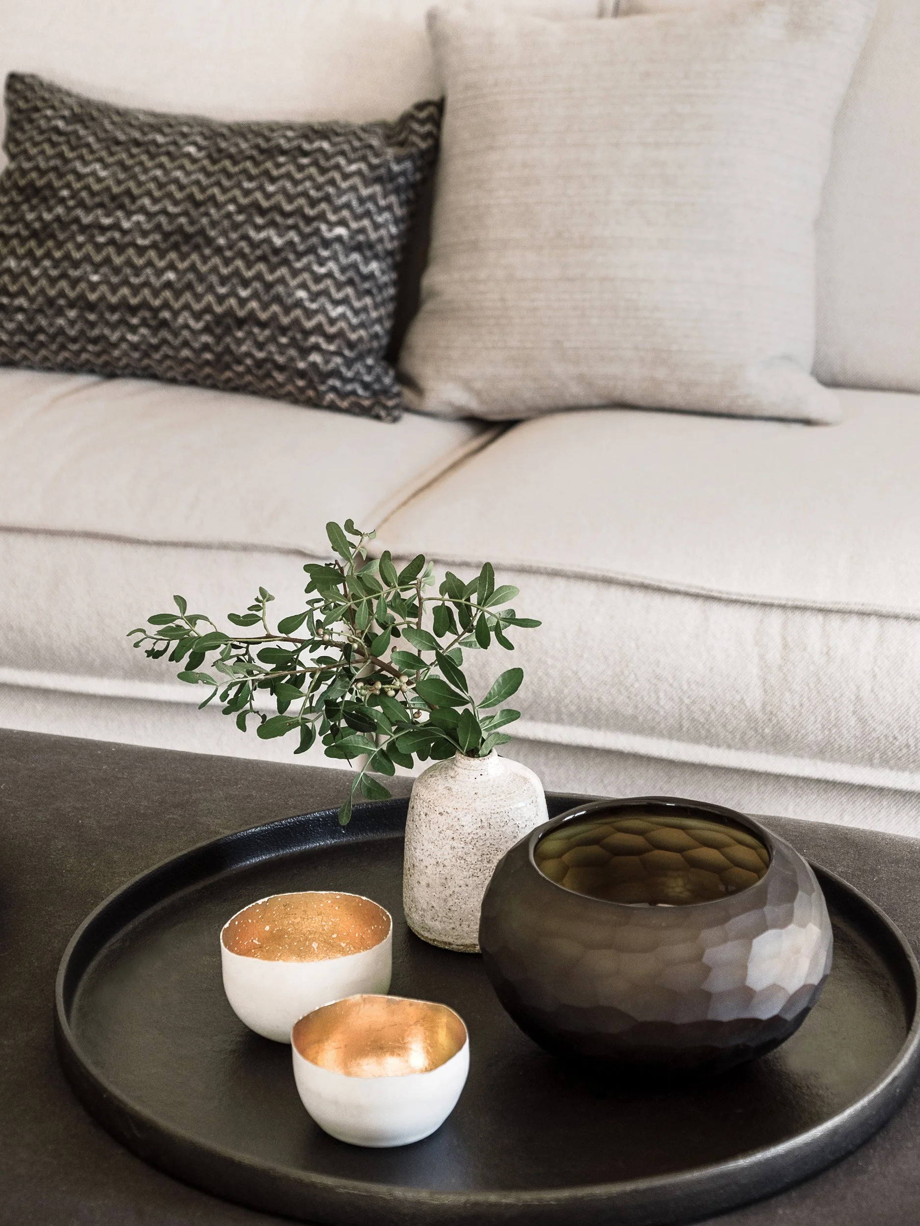 A black tray holding a small white bowl with gold interior, two more identical bowls, a large black faceted bowl, and a small white vase with green leaves, all on a dark surface in front of a beige sofa with patterned and plain cushions.