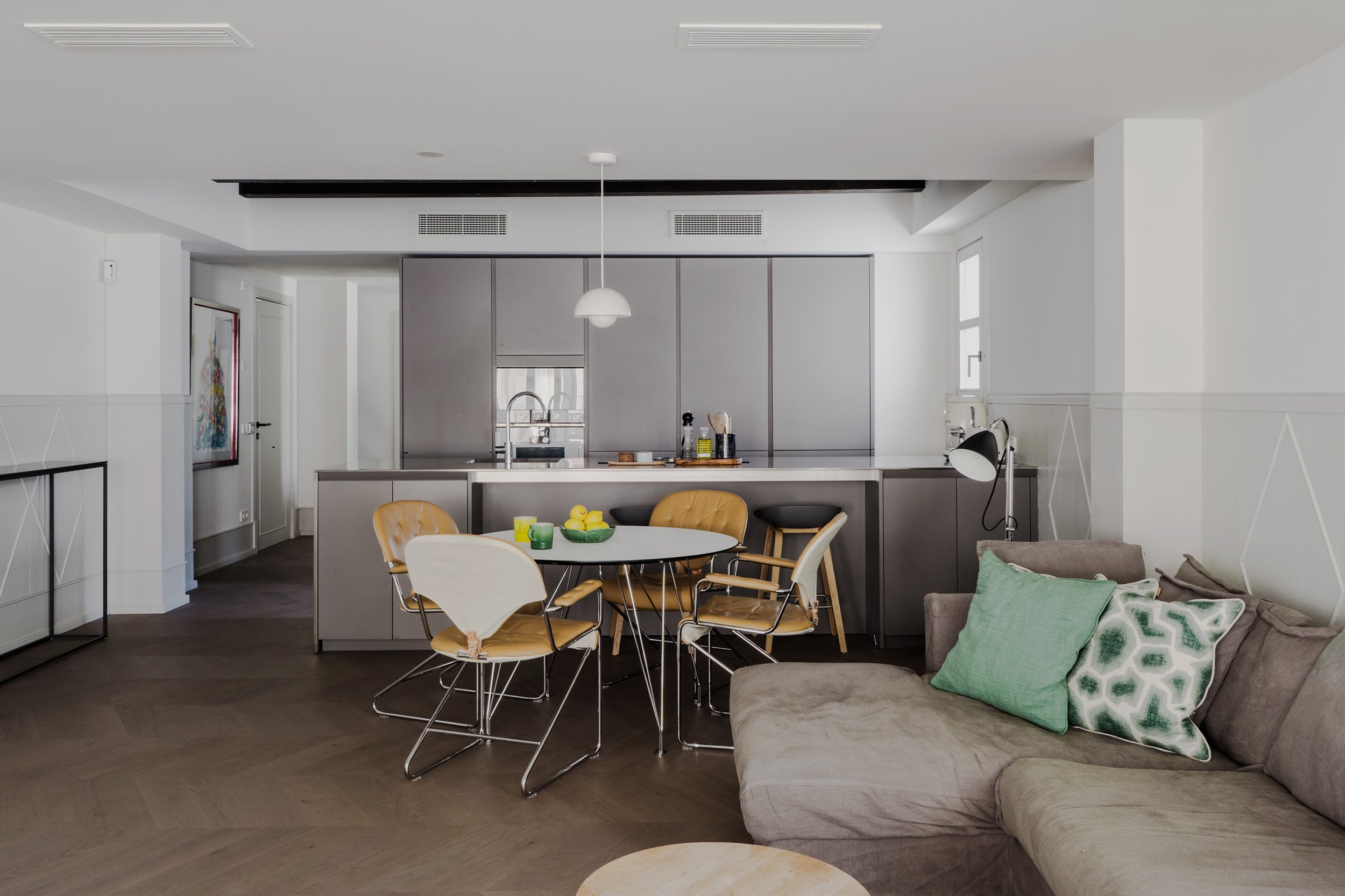 Open-concept living space with a kitchen, dining area, and living room. The kitchen features grey cabinetry, a sink, and modern appliances. The dining table has yellow and white chairs, and the living room has a beige sofa with green accent pillows. There are windows allowing natural light to brighten the space.