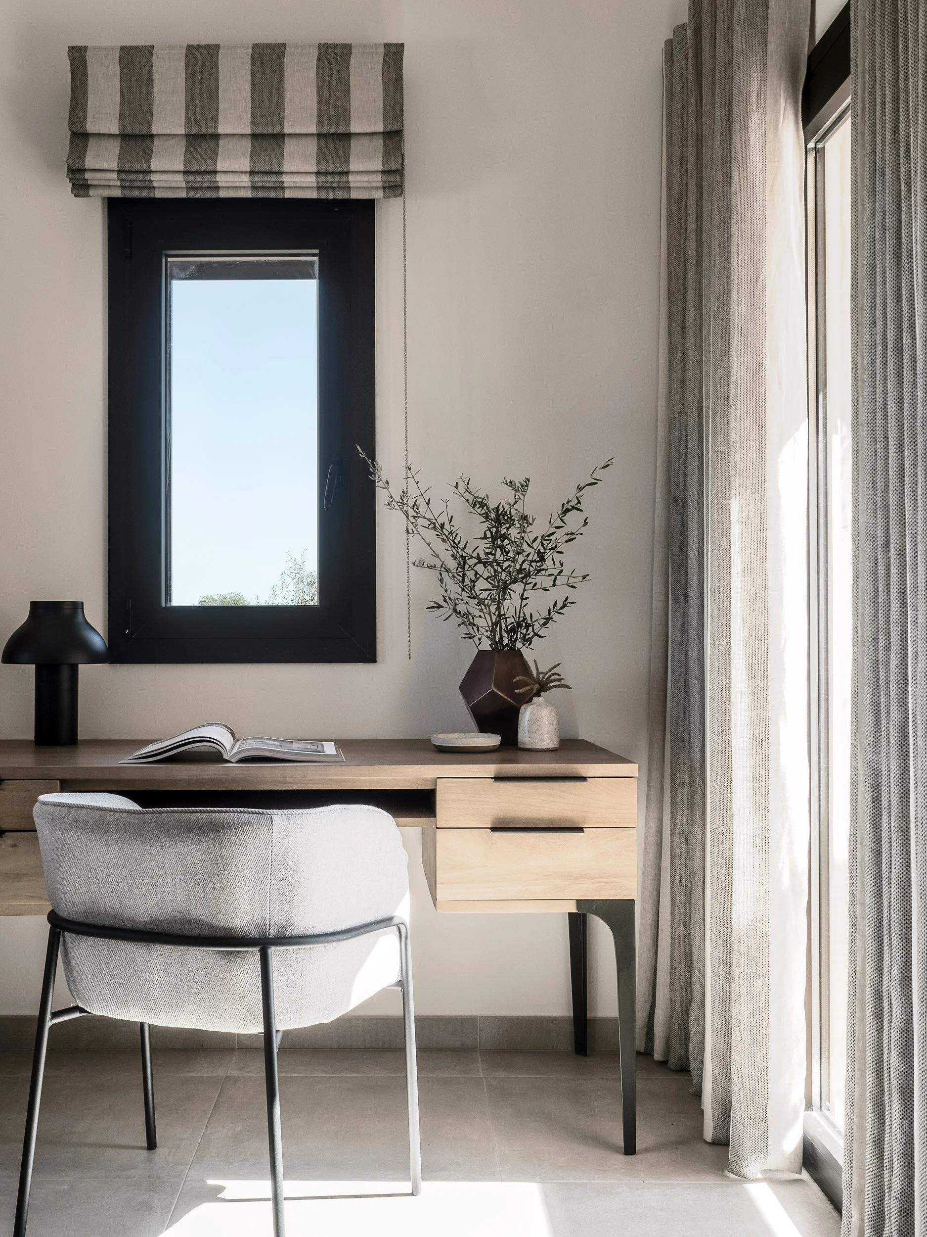 A modern desk area with a light wood desk, a gray upholstered chair, a black table lamp, an open magazine, a decorative vase with green branches, and a window with a striped gray and beige Roman shade and patterned curtains, allowing sunlight to brig
