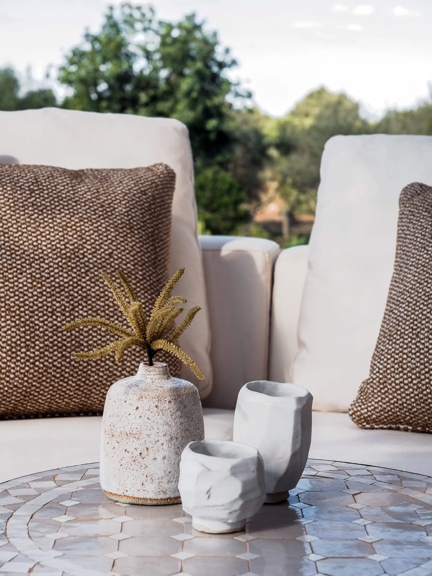 A beige outdoor sofa with brown and white patterned cushions, a small beige vase with a dried plant, and three small white marble or ceramic containers on a round tiled table, with trees and a partly cloudy sky in the background.