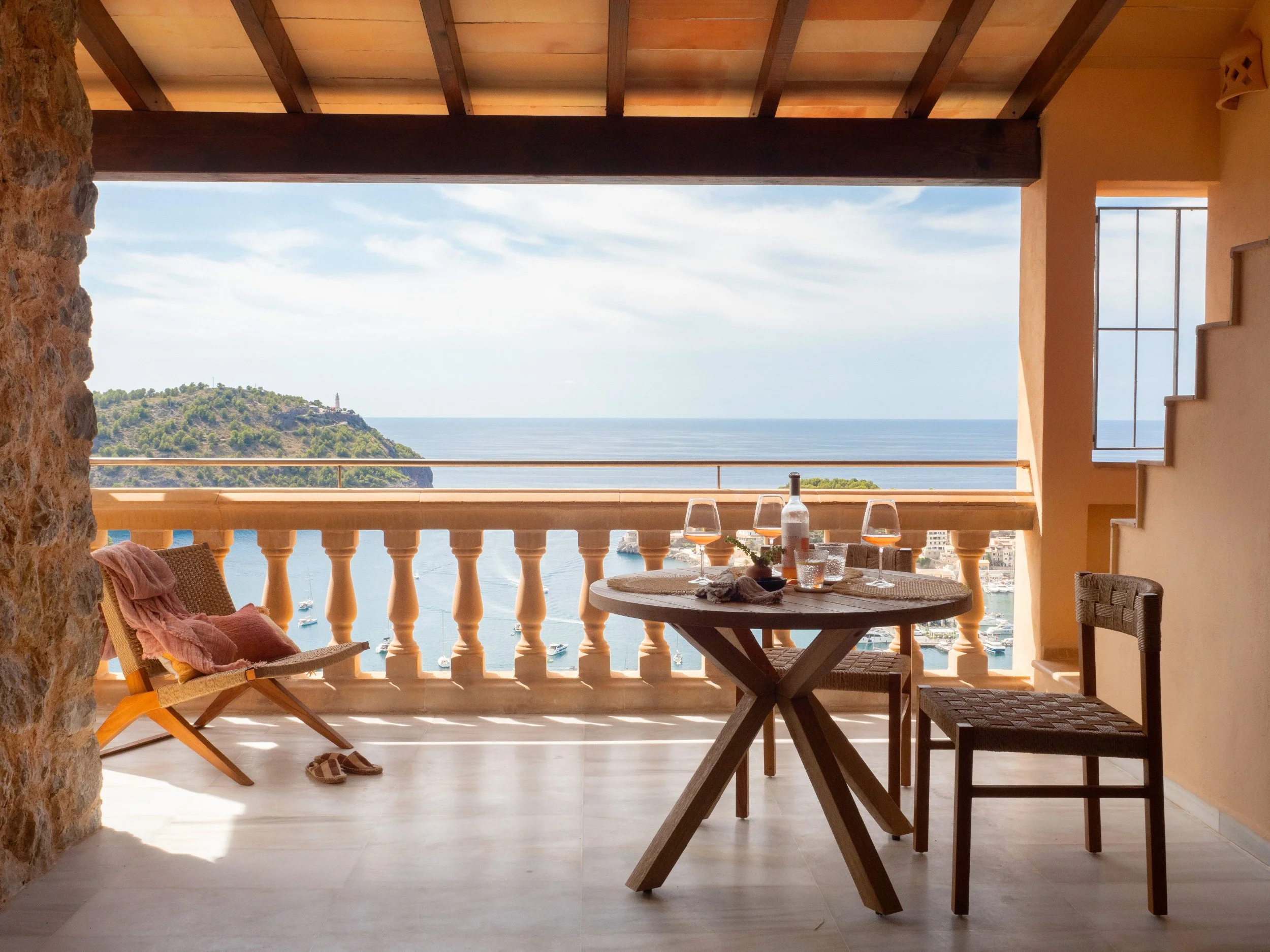 Balcony view overlooking a harbor with boats, a hill with greenery, and the ocean in the distance. The balcony has a round dining table with wine glasses and a bottle, two chairs, and a lounge chair with a towel.