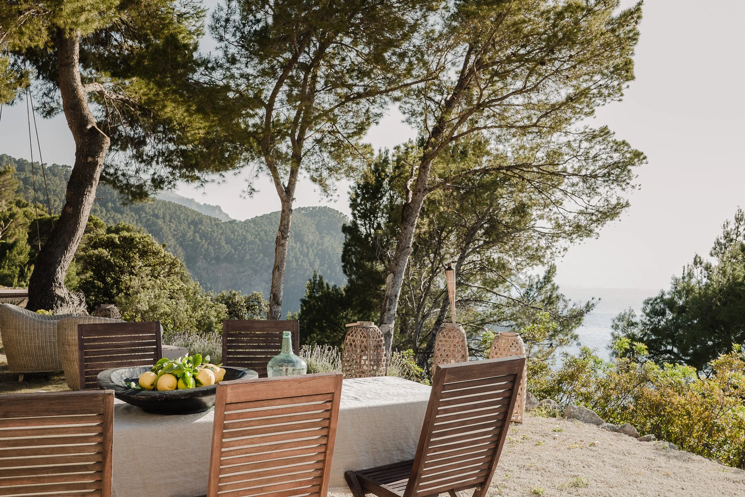Outdoor dining area with a table, chairs, and a fruit bowl, overlooking trees and mountains in the background.