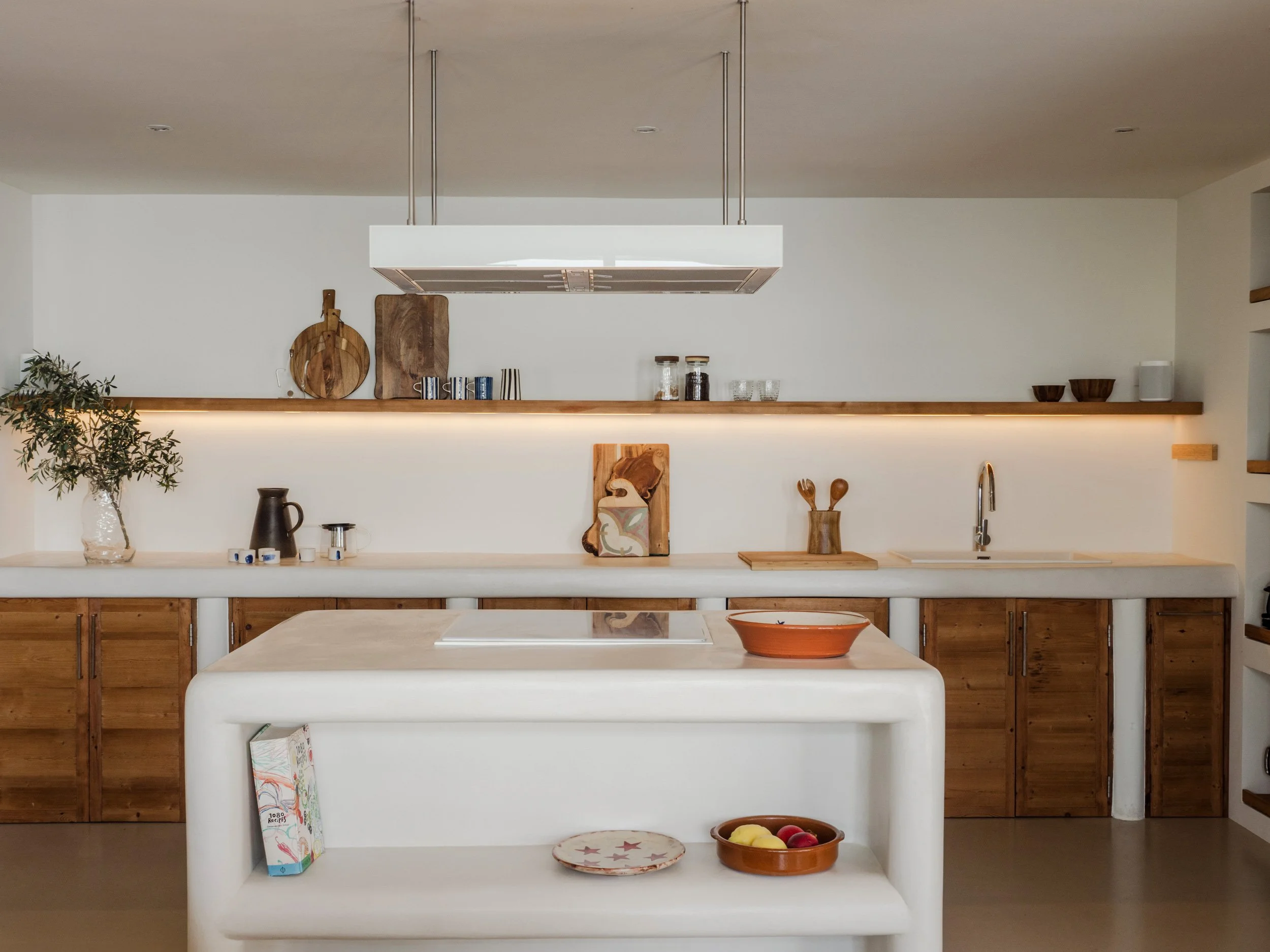 A kitchen with a white island and wooden cabinets, decorated with a potted plant, wooden cutting boards, jars, glasses, bowls, and a fruit basket, under a modern white pendant range hood.