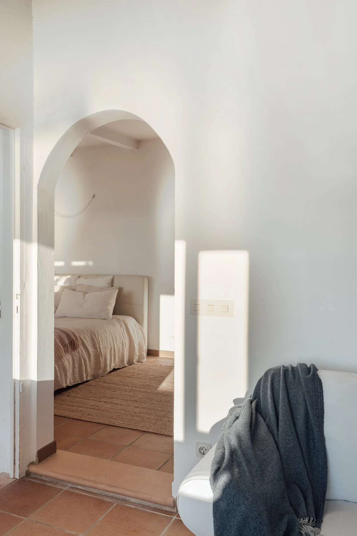 A view through a doorway into a bedroom with a bed covered in light-colored bedding. The room has a white headboard and beige pillows. Adjacent to the bed is a beige rug on a terracotta-tiled floor. To the right, part of a white sofa with a dark gray