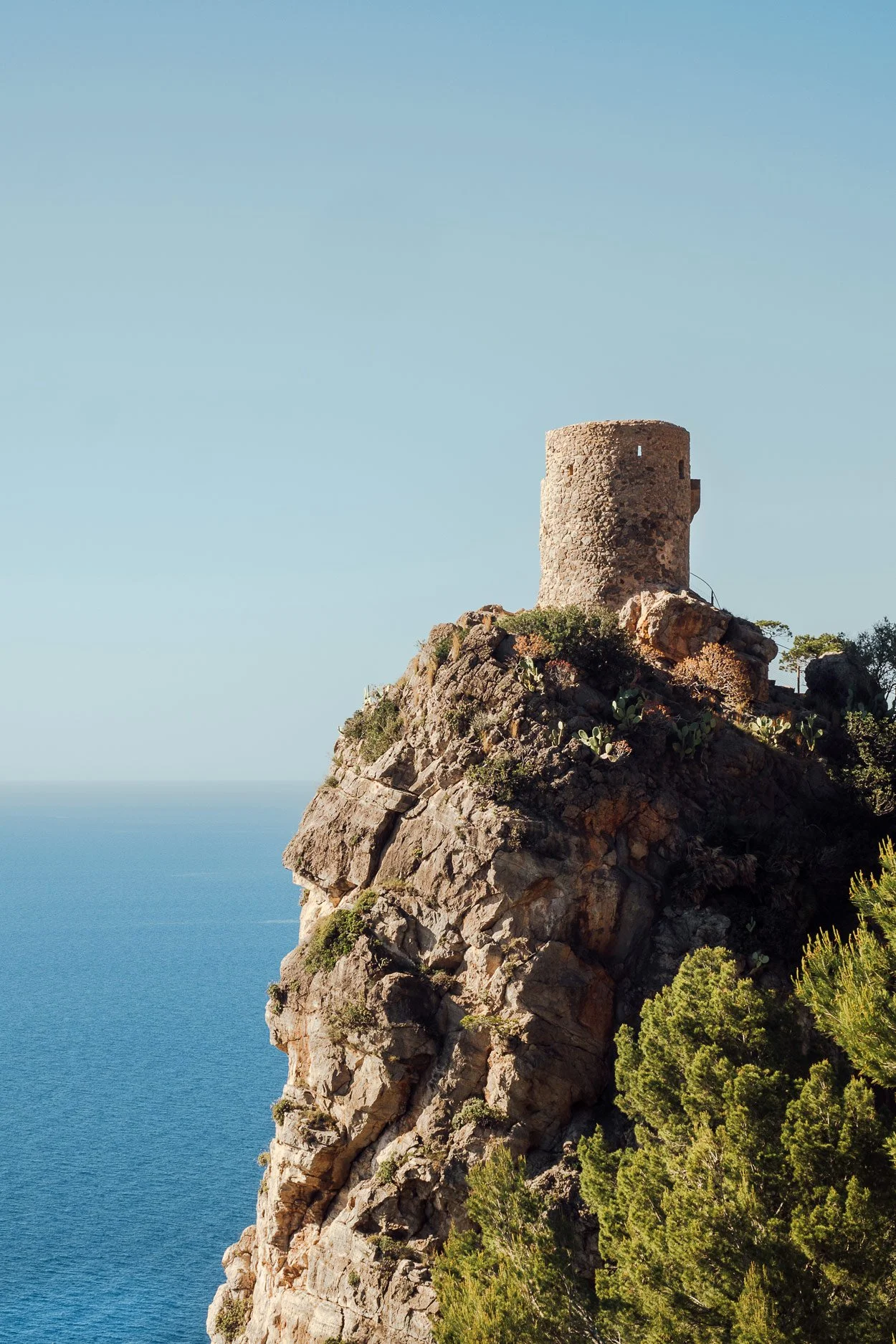 Ancient stone tower on a rocky cliff overlooking the sea with green vegetation and a clear blue sky.