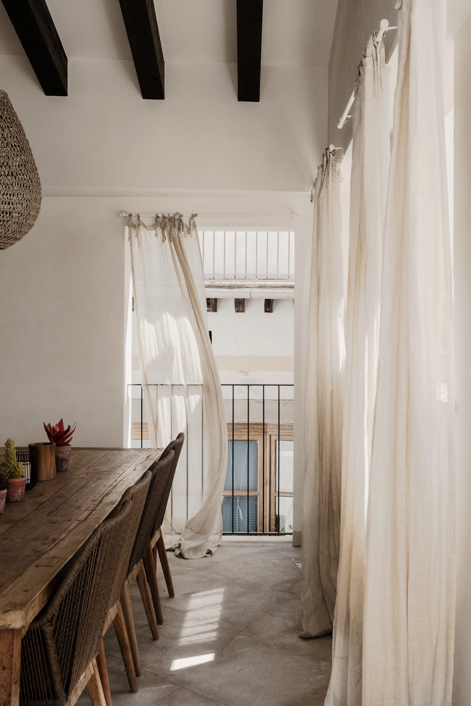 A cozy room with a dining table, chairs, sheer curtains, and a balcony with black metal railing and some exposed ceiling beams.