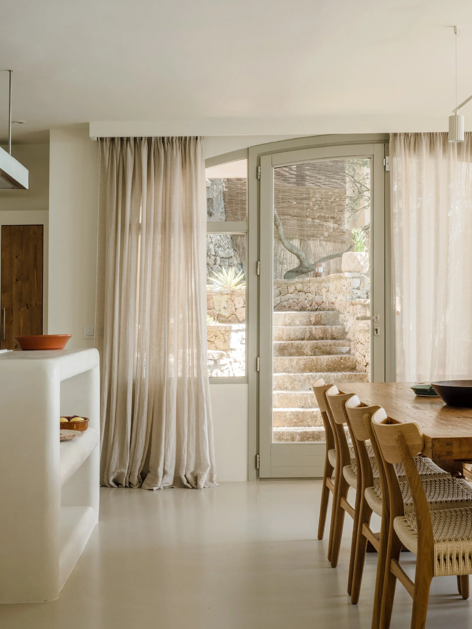 A bright living room with a door leading outside, beige curtains, a staircase outside, and a wooden dining table with chairs.
