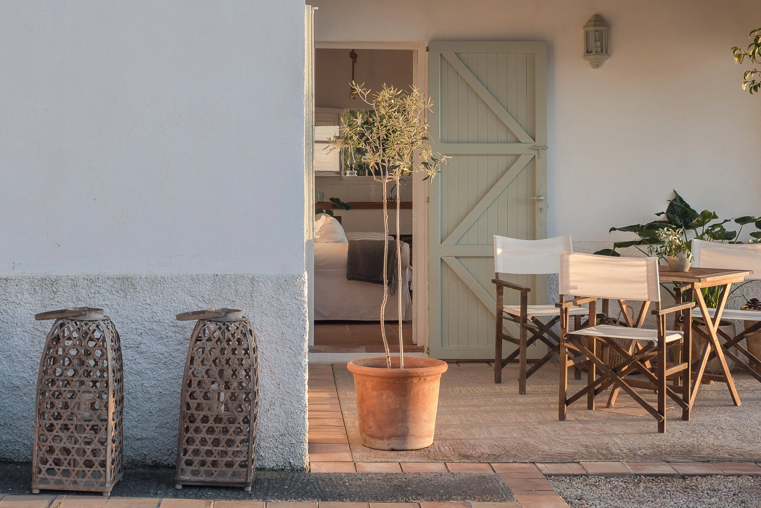 A cozy patio with outdoor chairs and a table, a potted tree, and decorative lanterns, with an open door leading into a bedroom with a bed and greenery inside.