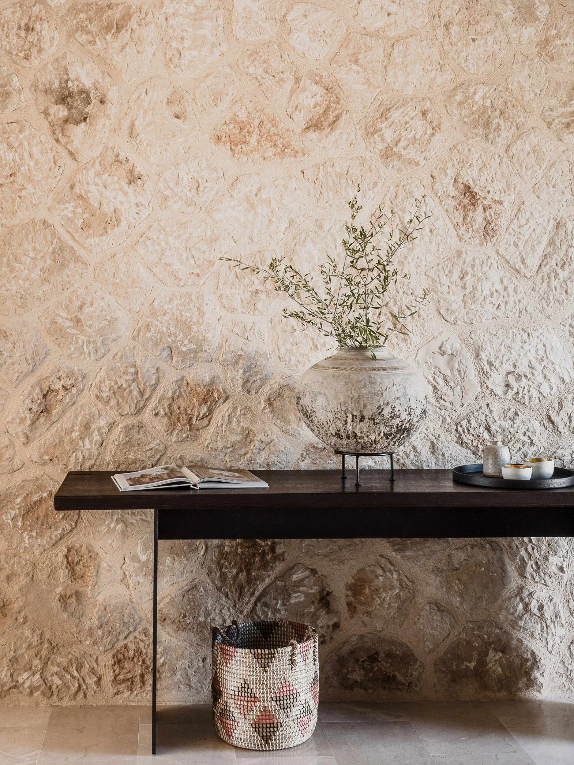 A dark wooden table against a beige textured stone wall with a large white and black ceramic vase holding green leafy branches, an open magazine, three small white and gold bowls, and a tall white container on a black tray.