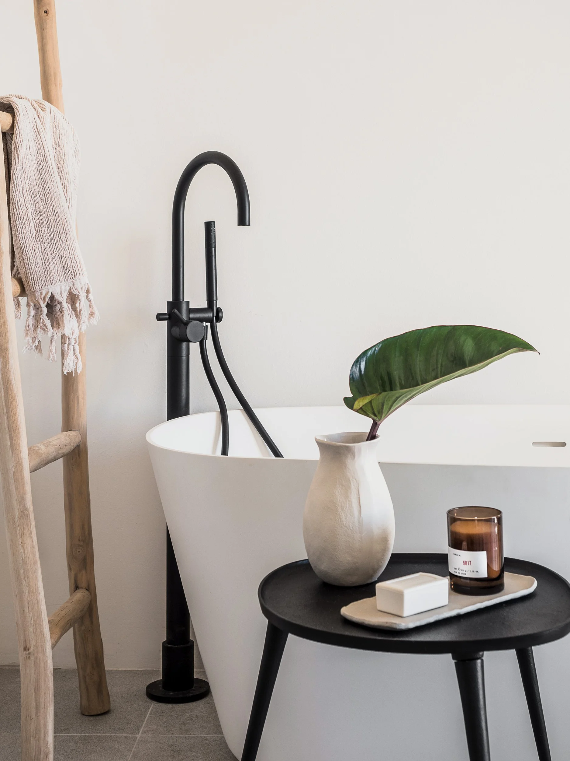 A bathroom scene featuring a white bathtub, a black faucet, a wooden towel rack with a beige towel, leafy green plant in a white vase, a small brown candle, and a tray with a white rectangular box and a small white item, all against a plain white wal