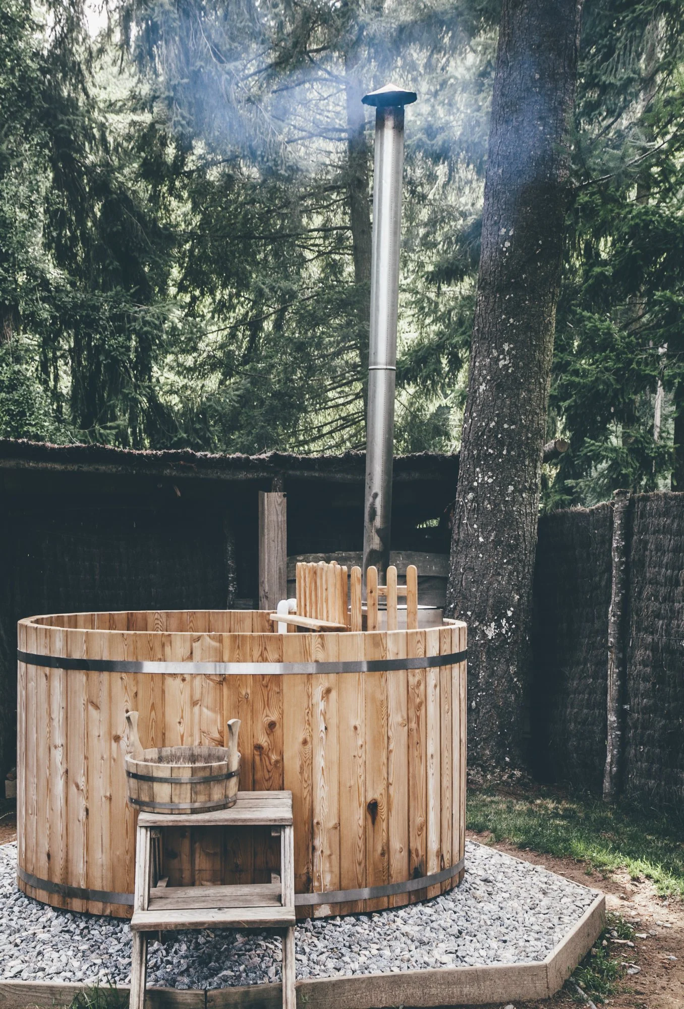 A wooden hot tub with a small step and a bucket on top, set in a backyard with pine trees and a black fence, with a tall metal chimney and some smoke rising.