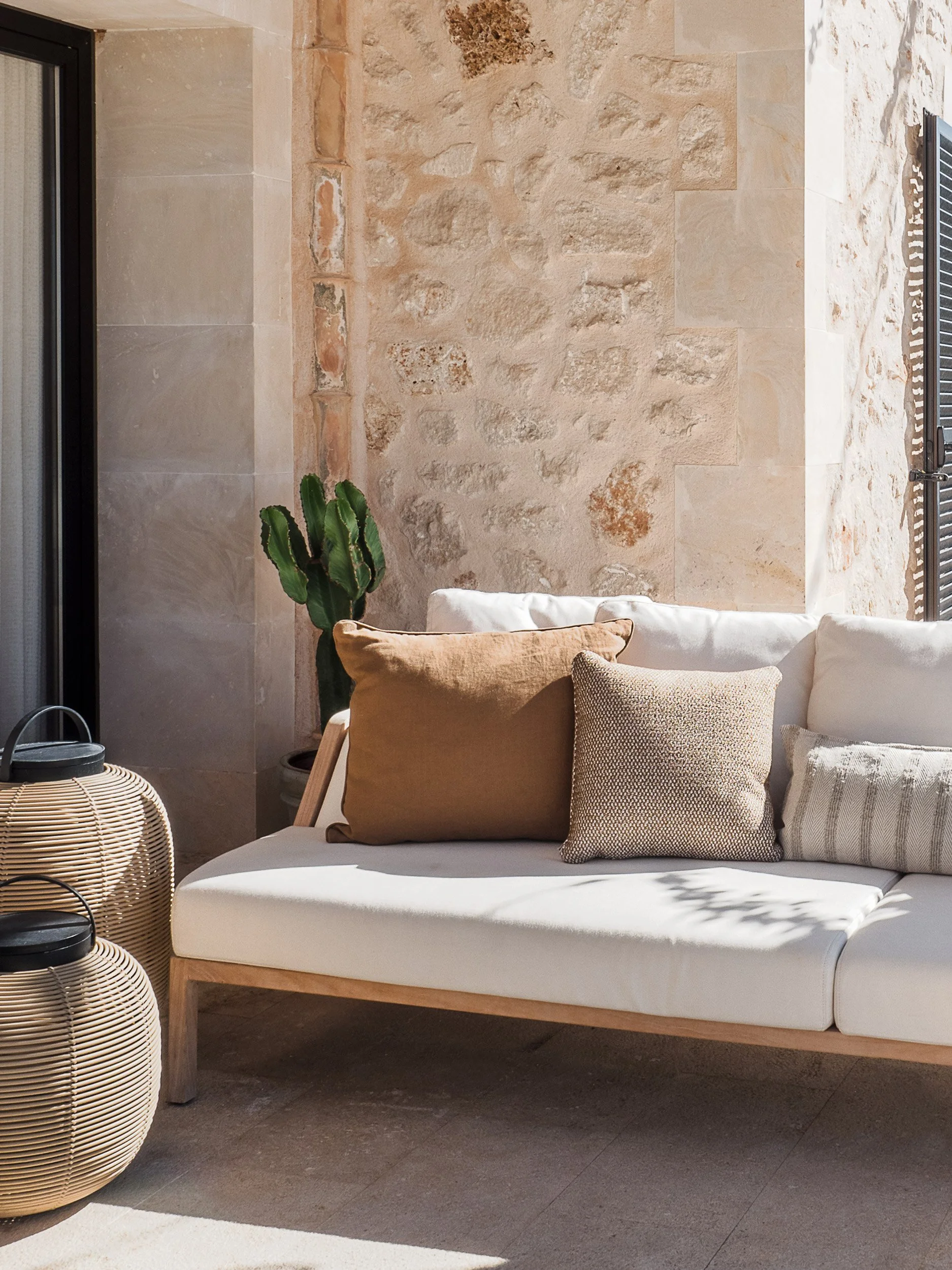 Outdoor seating area with a white cushioned sofa, beige pillows, a green cactus plant, and woven basket side table set against a stone wall and window with black shutters.