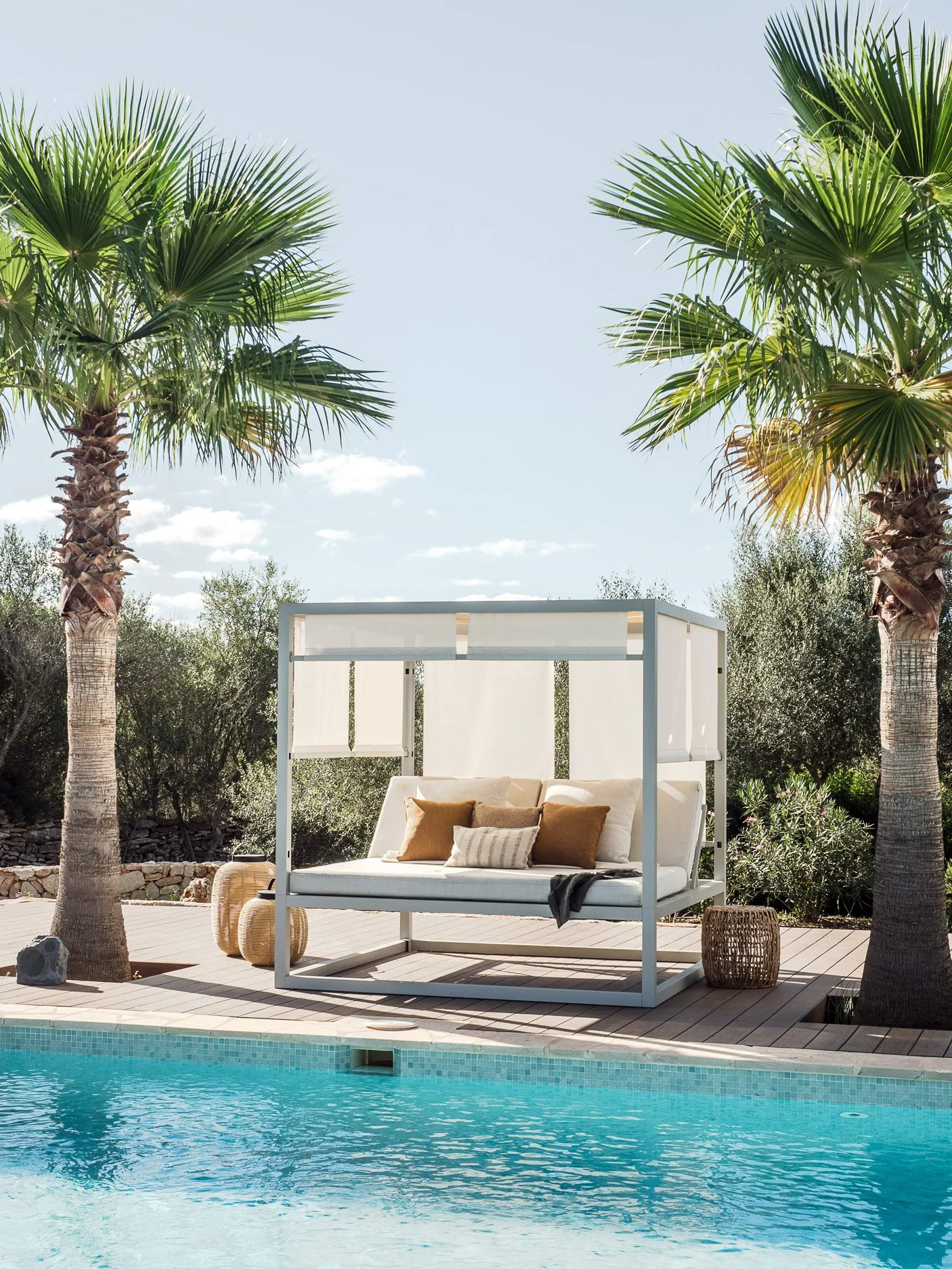 A poolside scene with a white canopy bed and beige pillows, surrounded by palm trees and outdoor decor, overlooking a swimming pool under a blue sky.