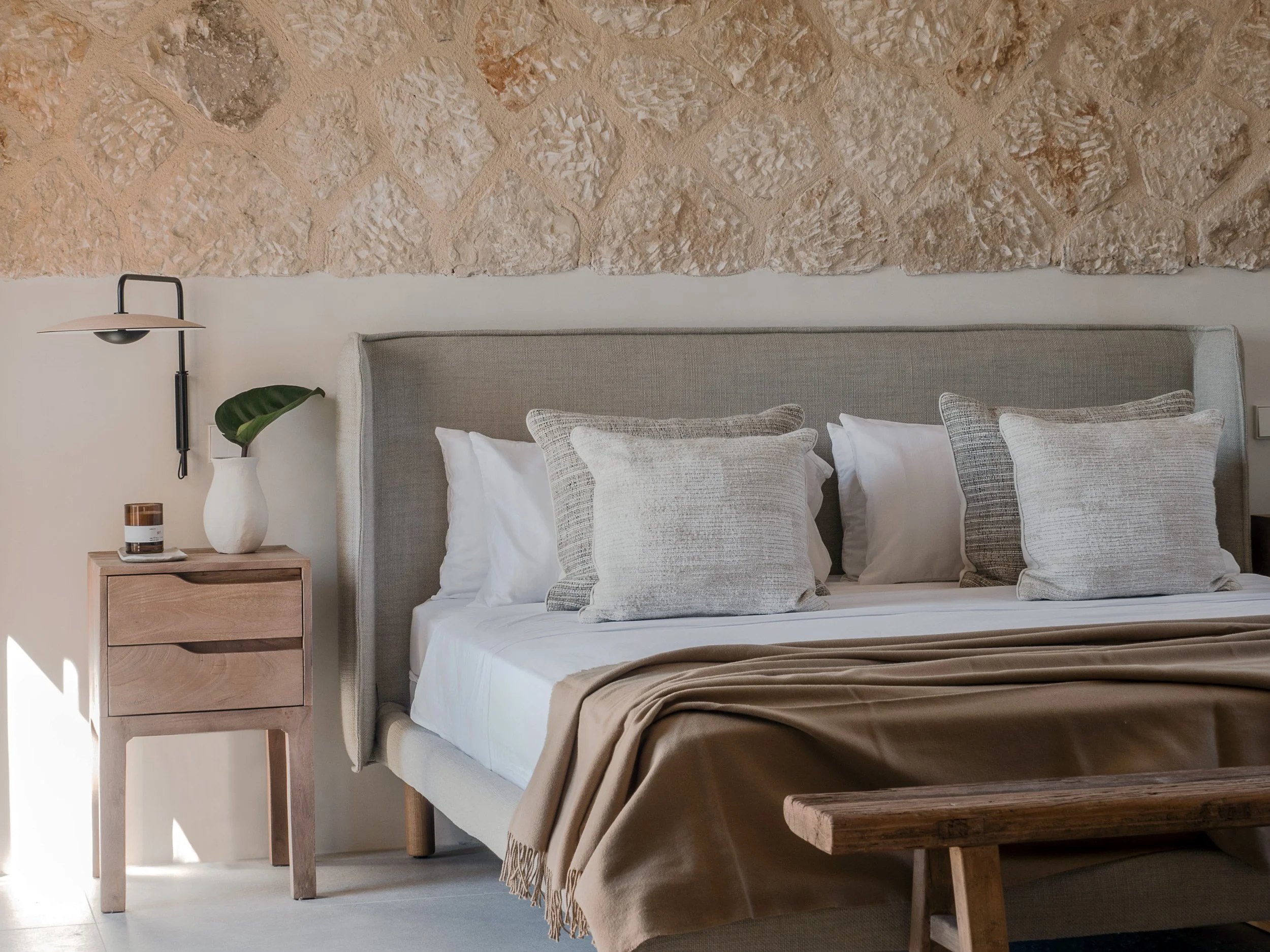 A bedroom with a beige upholstered bed, decorative pillows, a wooden nightstand with a white vase containing a large green leaf, a candle, and a modern black and white wall-mounted lamp. The wall behind the bed features an exposed stone accent wall.