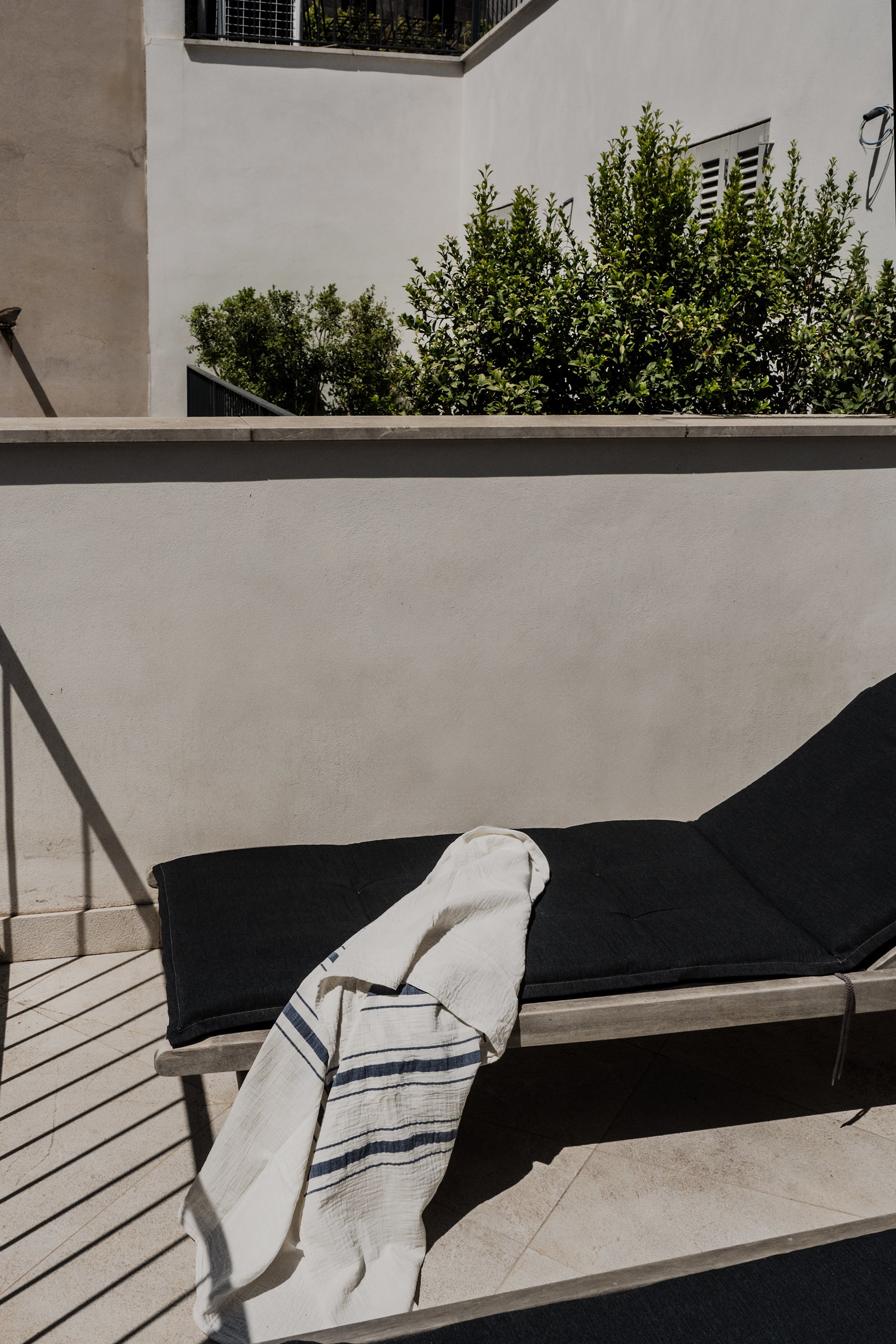 An outdoor patio scene featuring a black outdoor sofa with a white and blue striped towel draped over the arm and a white cloth on the seat, against a white wall with a railing, with greenery visible beyond the wall.